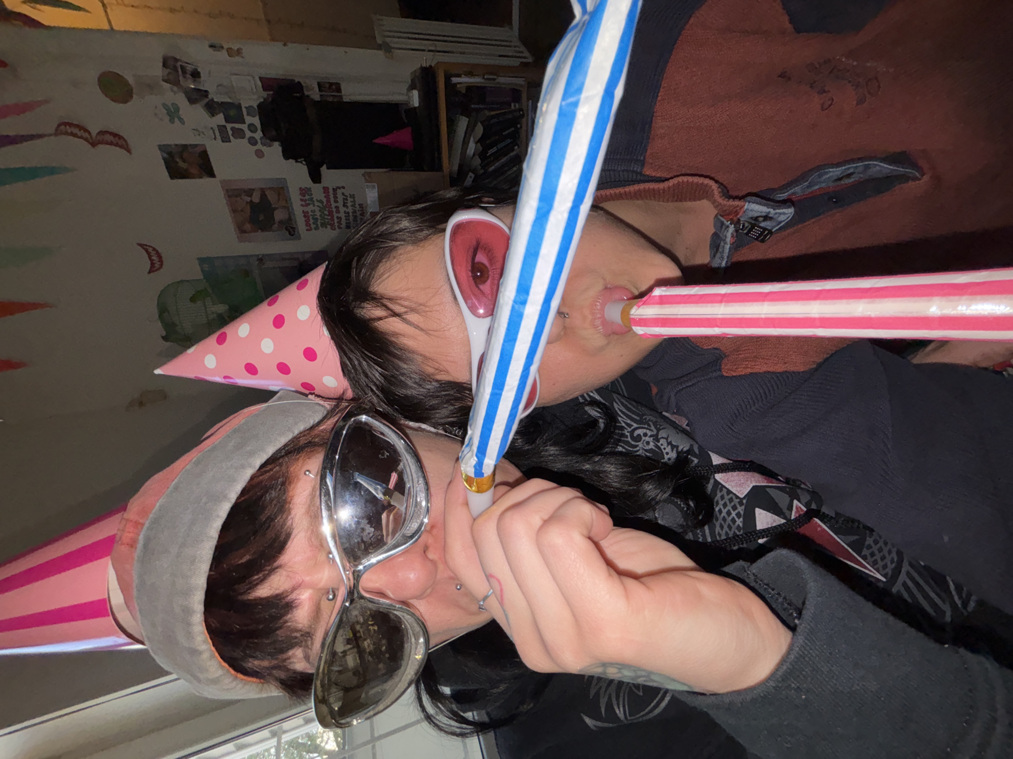 Two individuals are visible in an indoor setting. The person in the foreground is wearing a pink and grey party hat and large silver sunglasses and blowing a blue and white party horn. The other person, located in the background, is wearing a pink and white party horn and white sunglasses with red lenses. Both people are blowing party horns. The background includes a wall with various posters and decorations. It is not possible to ascertain the location as the city of Sofia, Bulgaria from this image.