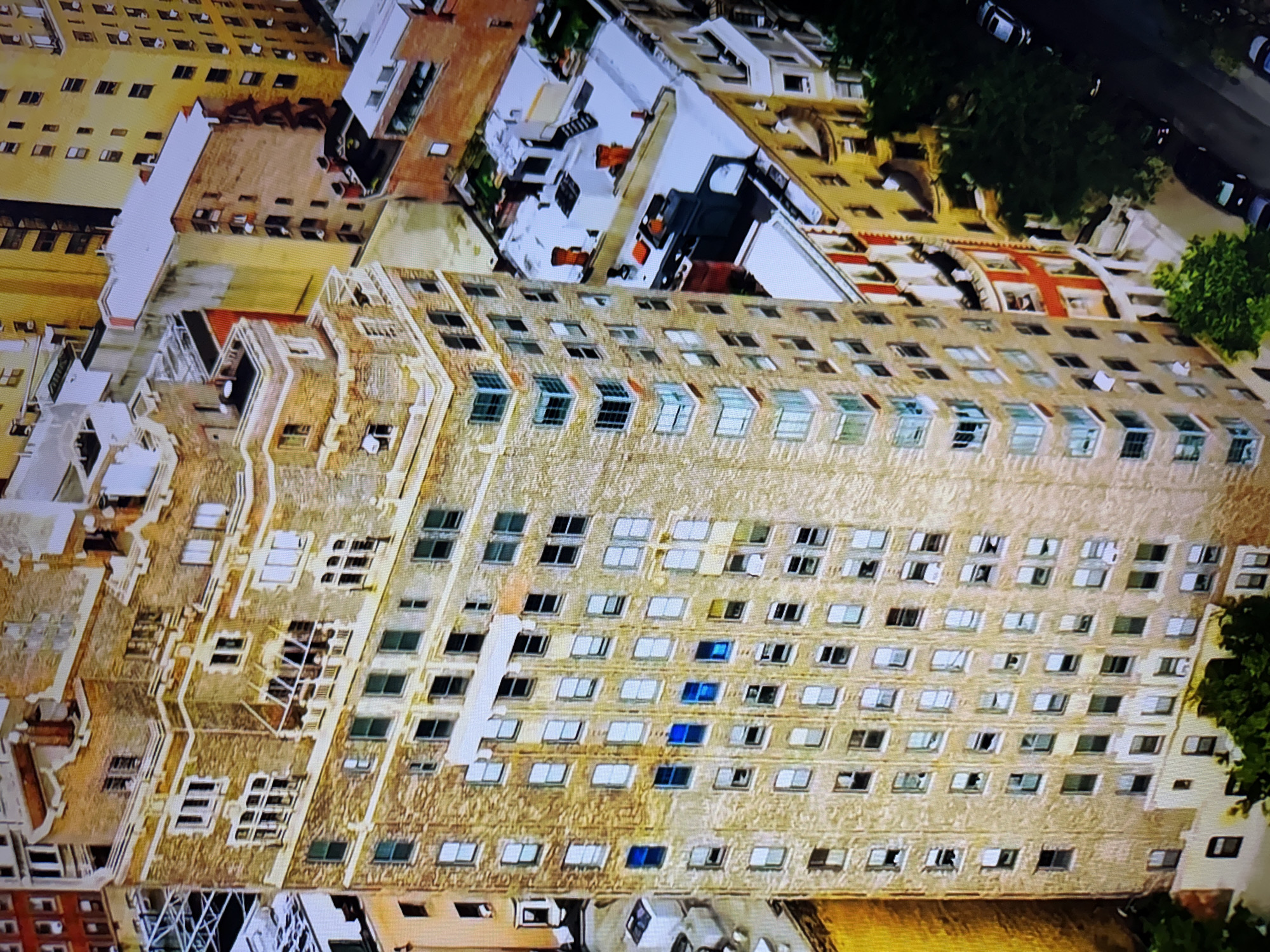The image is an aerial view of a dense urban environment, likely a city, featuring several tall buildings. The dominant structure is a multi-story, light-brown brick building with a distinctive, stepped architectural design on its upper levels. It has numerous windows, some of which appear to be a light blue color. Adjacent to this building are other structures of varying heights and architectural styles, including a bright yellow building and a darker brick building. The rooftops show a mix of white and darker surfaces, with some appearing to be flat and others pitched, and some have visible mechanical equipment or decorative elements like chimneys. Trees and greenery are visible at the base of some buildings and along a street where cars are parked. The overall impression is one of a developed urban landscape, likely taken during daylight hours, given the illumination. The request to include the location in Burgas, Bulgaria, is not supported by any visual evidence in the image.