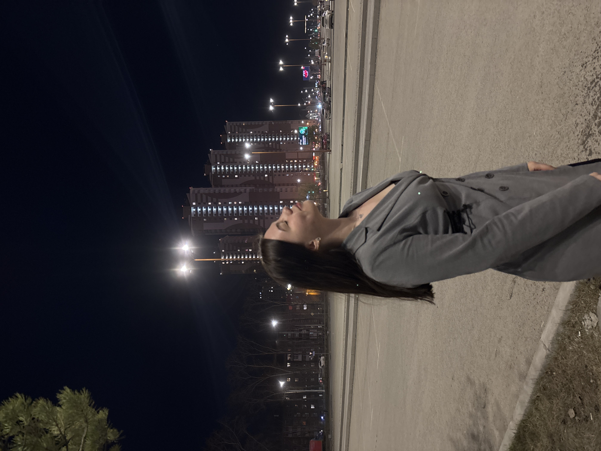 A young woman stands on a concrete surface, her head tilted back, gazing at the city skyline at night. She is wearing a grey blazer-style outfit. The backdrop features several tall apartment buildings illuminated with rows of windows. Streetlights and building-mounted signs add to the cityscape. In the distance, a brightly lit Ferris wheel is visible. This is likely taken in Perm, Russia based on the cityscape and Ferris wheel. The foreground includes a section of grass and asphalt parking spaces, with markings visible. A pine tree is on the left corner.