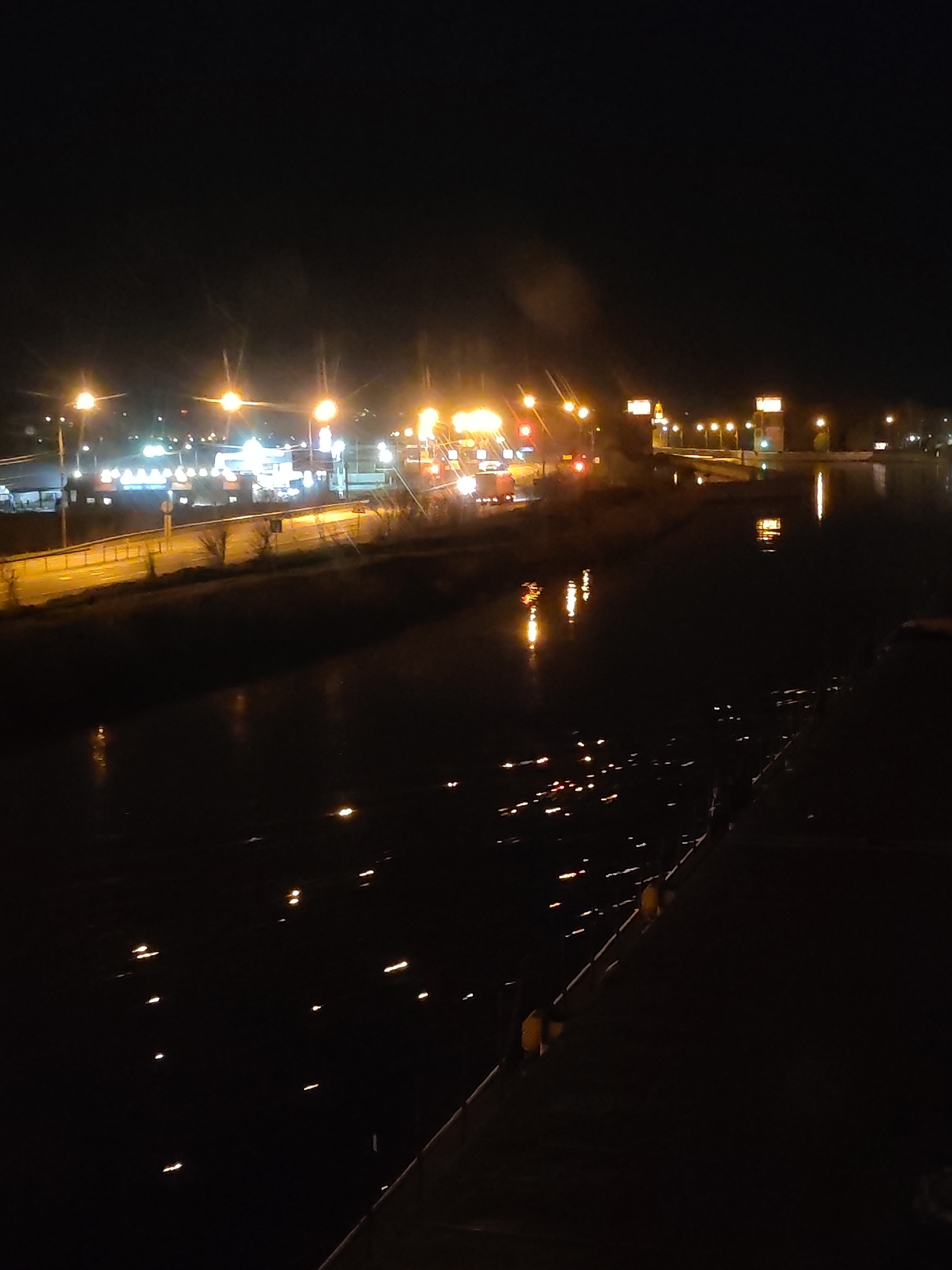 The image captures a night scene in Yakhroma, Russia, featuring a body of water reflecting city lights. The water appears to be a river or canal, with faint reflections suggesting the presence of streetlights and buildings along its banks. On the far side of the water, there is a road with streetlights illuminating the pavement and possible traffic. The overall environment is dark with the main light source coming from city lights and their reflection on the water.