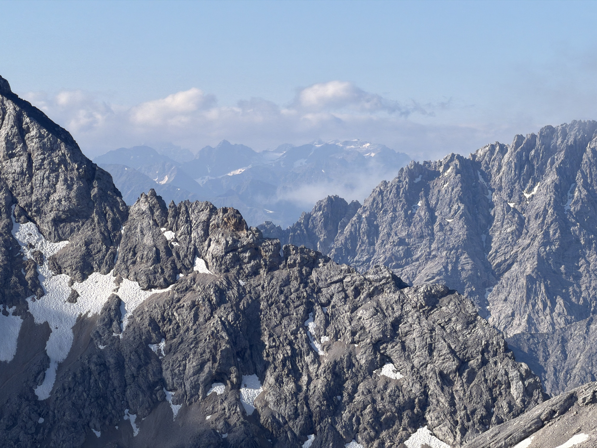 The image depicts a panoramic view of a rugged mountain range under a clear blue sky with scattered white clouds. The foreground features a steeply angled, rocky mountain slope with patches of snow. The middle ground shows more jagged mountain peaks, also with snow, fading into a hazy, distant mountain landscape. The scene is a natural mountain environment, typical of the Bavarian Alps, and is likely captured from a high vantage point, possibly a mountain peak or a viewpoint in the vicinity of Garmisch-Partenkirchen, Germany, known for its alpine scenery. There are no people or man-made structures visible in the image, focusing solely on the geological formations and atmospheric conditions of the mountains.