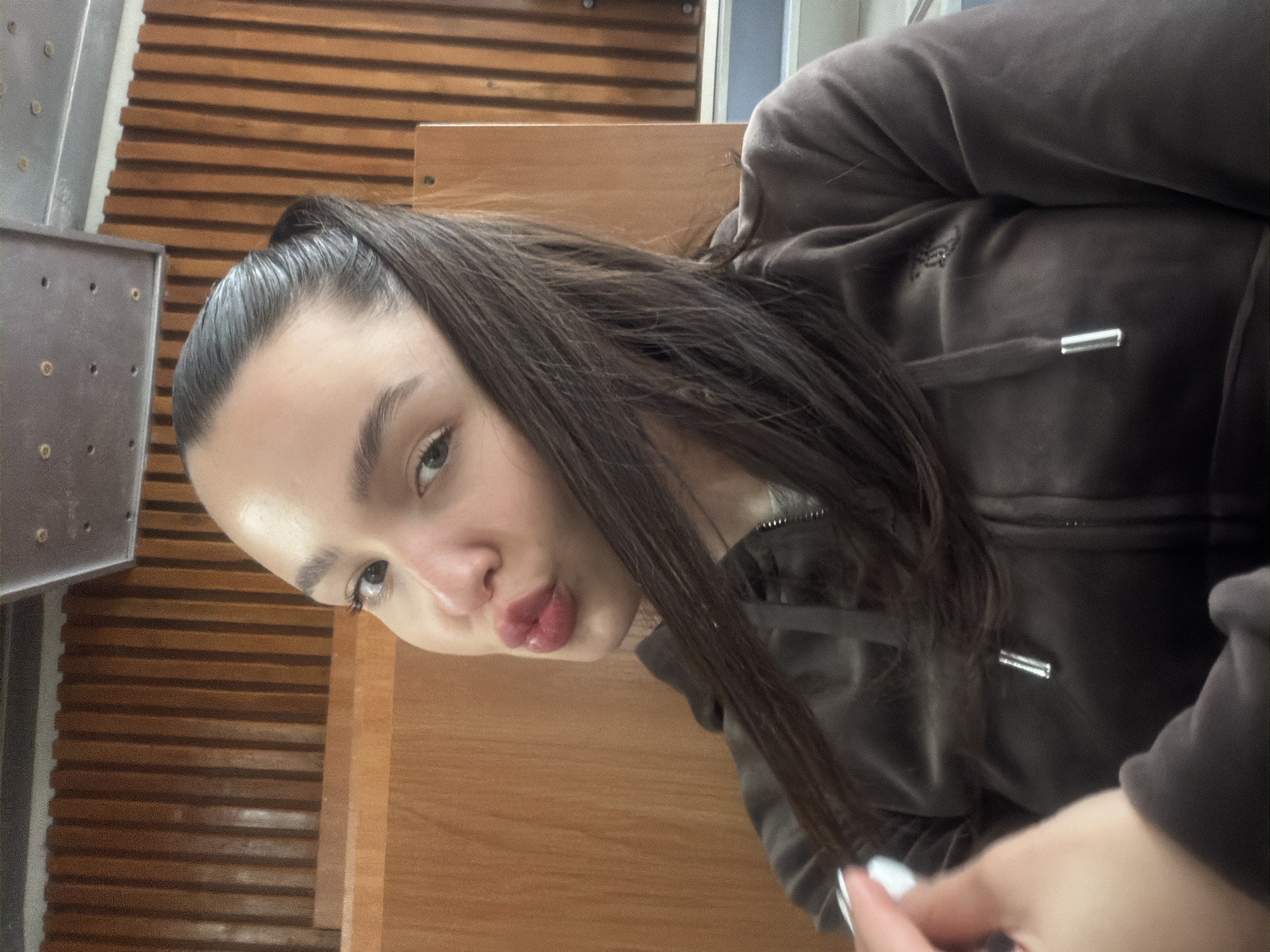A young woman is captured in a tilted selfie. She has long, dark brown hair, pulled back into a high ponytail, with some strands falling forward. Her light eyes are directed towards the camera, and her lips are pursed in a prominent pout, tinted with a reddish-pink color. She is wearing a dark brown, possibly velour or velvet, hooded zip-up jacket. A distinctive, embroidered crown-like logo is visible on the left chest area of her jacket, and a matching drawstring with silver tips extends from the hood. A light-colored nail polish is visible on her fingernails in the bottom right corner. The background features a patterned wooden surface with horizontal slats, suggesting interior paneling or furniture. To the left, a portion of a gray, possibly metallic, structure with circular indentations is visible. The scene appears to be indoors, likely in Sofia, Bulgaria.
