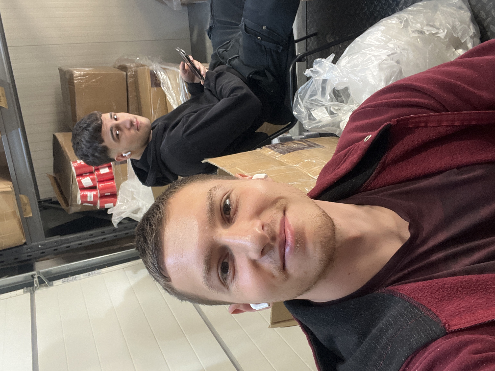 Two men are present in what appears to be a storage or warehouse environment. The man in the foreground is taking a selfie. He is wearing a dark red hooded sweatshirt over a dark top and has an earpiece in his ear. The second man is in the background, sitting in a chair with his legs propped up, looking towards the camera while holding a phone. He is wearing a black hooded sweatshirt and dark pants. There are boxes and packaging materials visible, suggesting a storage or shipping area. The location of Burgas, Bulgaria cannot be determined from the image.