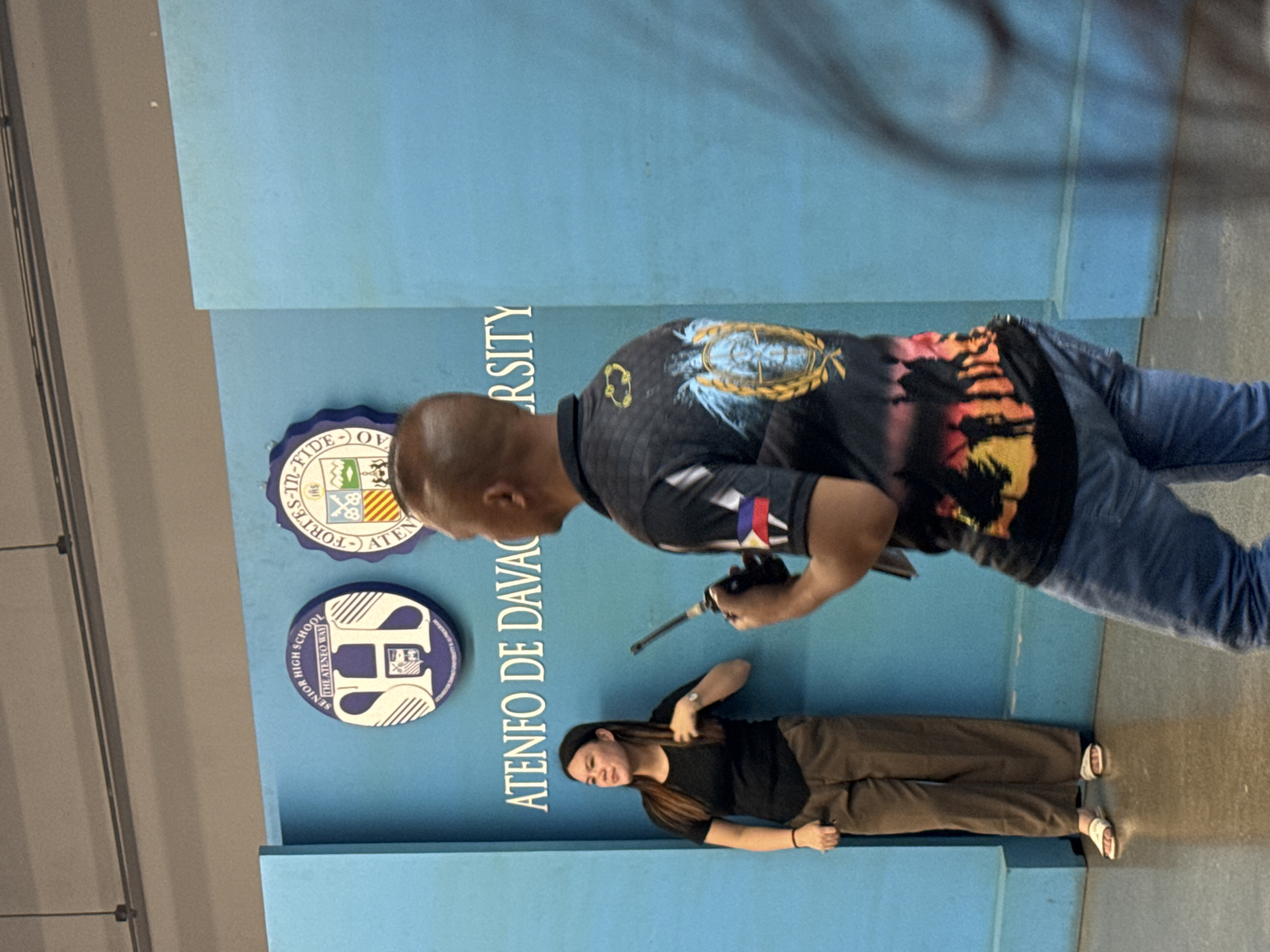 In an indoor setting with a vibrant blue backdrop, a woman stands to the left, leaning against a blue partition. She is wearing a black t-shirt and brown trousers, with white sandals on her feet. Her gaze is directed upwards and to her left. To her right, a man is partially visible from the back, wearing a dark-colored t-shirt with graphics on it and blue jeans. He is holding a black object, possibly a communication device, in his right hand. The blue backdrop features two circular logos and text that reads "ATENEO DE DAVAO". One of the logos is for the "SENIOR HIGH SCHOOL THE ATENEO WAY", and the other is a seal with the text "FORTES-IN-FIDE". The overall scene suggests an event or gathering at Ateneo de Davao University.