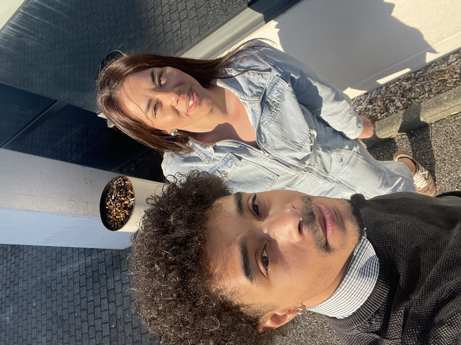 This image captures two individuals, a male and a female, positioned outdoors in Montigny-le-Bretonneux, France, under direct sunlight. The male, in the left foreground, exhibits dark, curly hair, a mustache, and a short beard. He is dressed in a black knitted sweater worn over a white and blue striped collared shirt, with a silver hoop earring visible in his left ear, and he is looking directly at the camera. To his right and slightly behind, the female has straight, dark hair and is smiling towards the camera. She wears a light-wash denim full-zip jumpsuit and a silver stud earring in her left ear. Her feet are visible, clad in light brown lace-up shoes with white soles. In the background, behind the male, a white cylindrical ashtray overflowing with numerous cigarette butts is present. The ground features dark grey interlocking bricks transitioning to a concrete surface with a raised curb. A light-colored building column and a dark, reflective building facade are visible in the background. Scattered cigarette butts and small pebbles are observed on the ground along the curb.