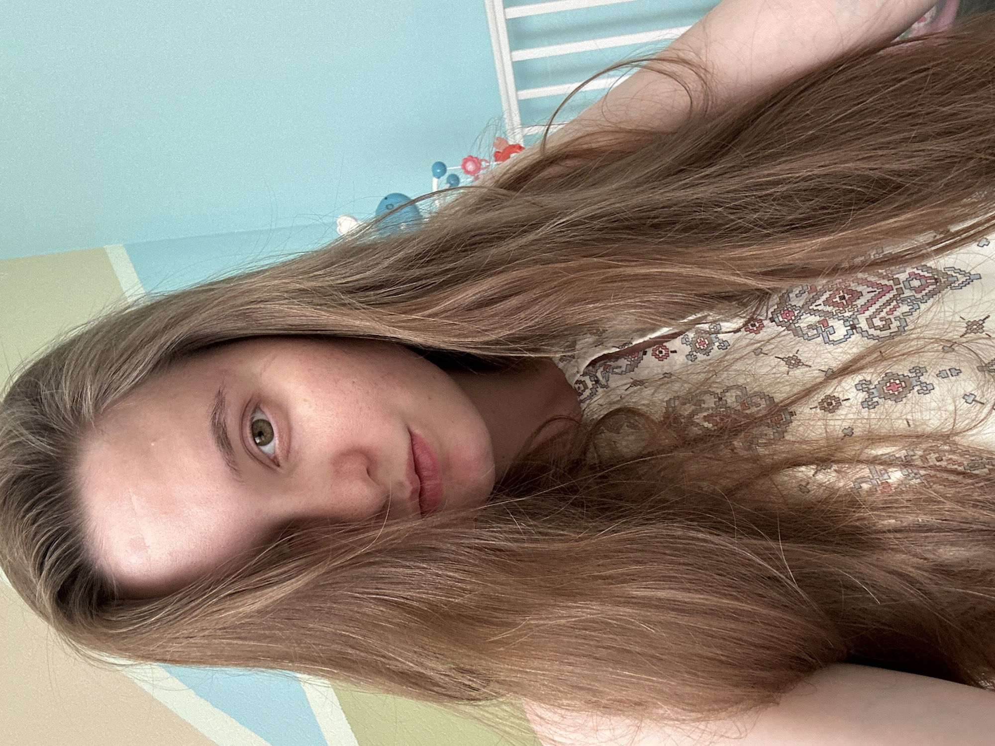 The image is a selfie of a young Caucasian woman with long, light brown hair that covers part of her face. She has green eyes and appears to be wearing a light-colored top with a patterned design. The background is a room with a light blue wall and a crib with a mobile hanging above it. There is also a wall with a geometric pattern in the background. The scene appears to be indoors, likely in a residential setting. There is no information provided to determine the exact location in the city of Noviy, Russia.