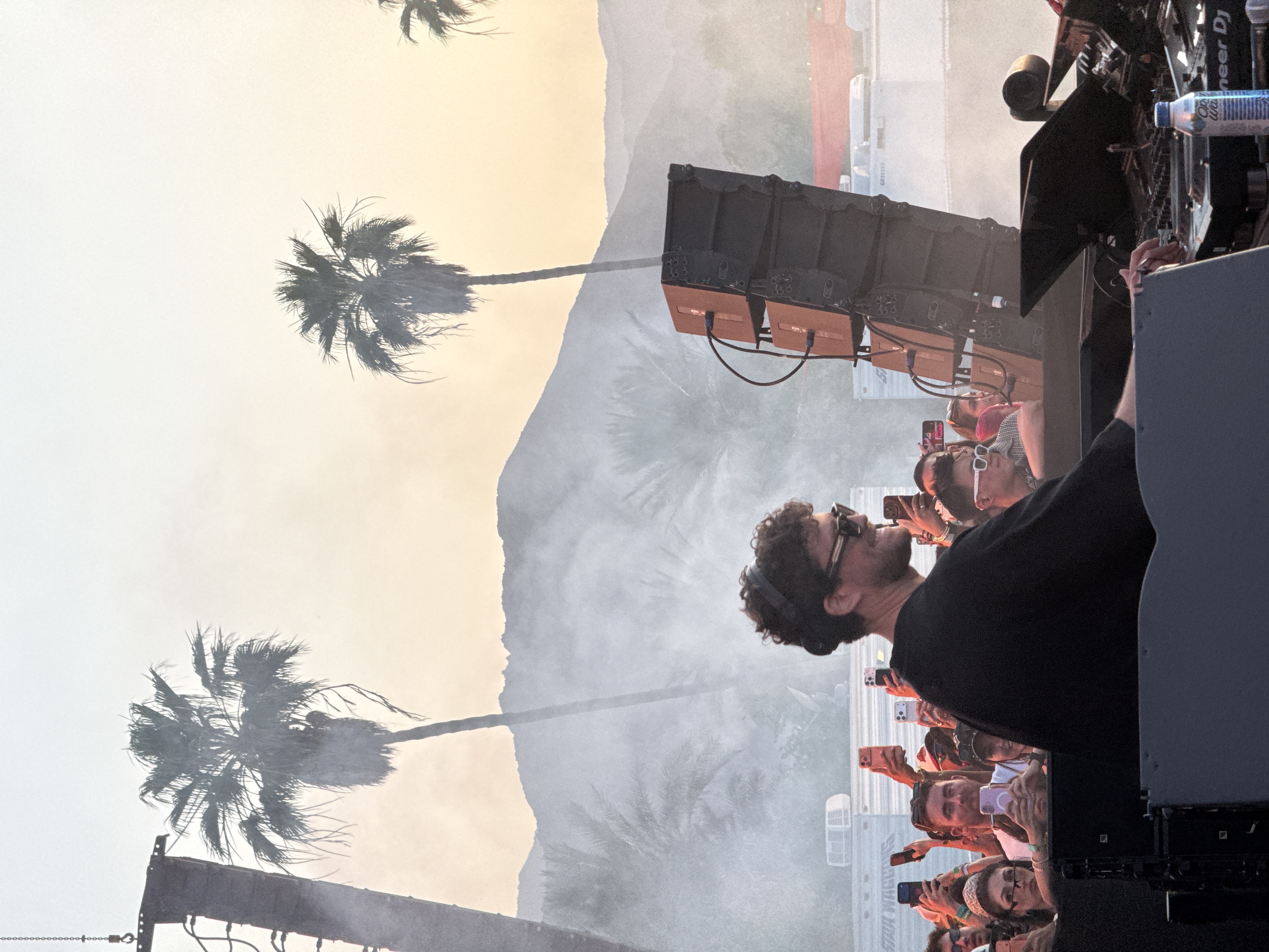 The image depicts an outdoor music event, likely a festival, taking place at dusk or early evening in a desert environment. In the foreground, a male DJ is positioned at a mixing console, seen from a side profile. He has curly hair, wears sunglasses and headphones, and is dressed in a black long-sleeved shirt, smiling towards the left side of the frame. His right hand is on the console, suggesting active mixing.
Behind and to the left of the DJ, a lively crowd of attendees is visible, with many people holding up smartphones to capture the performance. Their faces show engagement and excitement. To the DJ's right, a tall stack of professional audio speakers is set up.
The background features several prominent palm trees, silhouetted against a hazy, warm-toned sky that transitions from light yellow to a pale orange, indicative of twilight. Distant mountains are visible through a layer of atmospheric haze or stage smoke, adding to the desert setting. On the DJ's console, a white plastic water bottle with a blue label is visible, bearing the text "Old Water". Part of the text "Pioneer DJ" is also discernible on the equipment. The overall scene conveys an energetic and immersive concert atmosphere.
