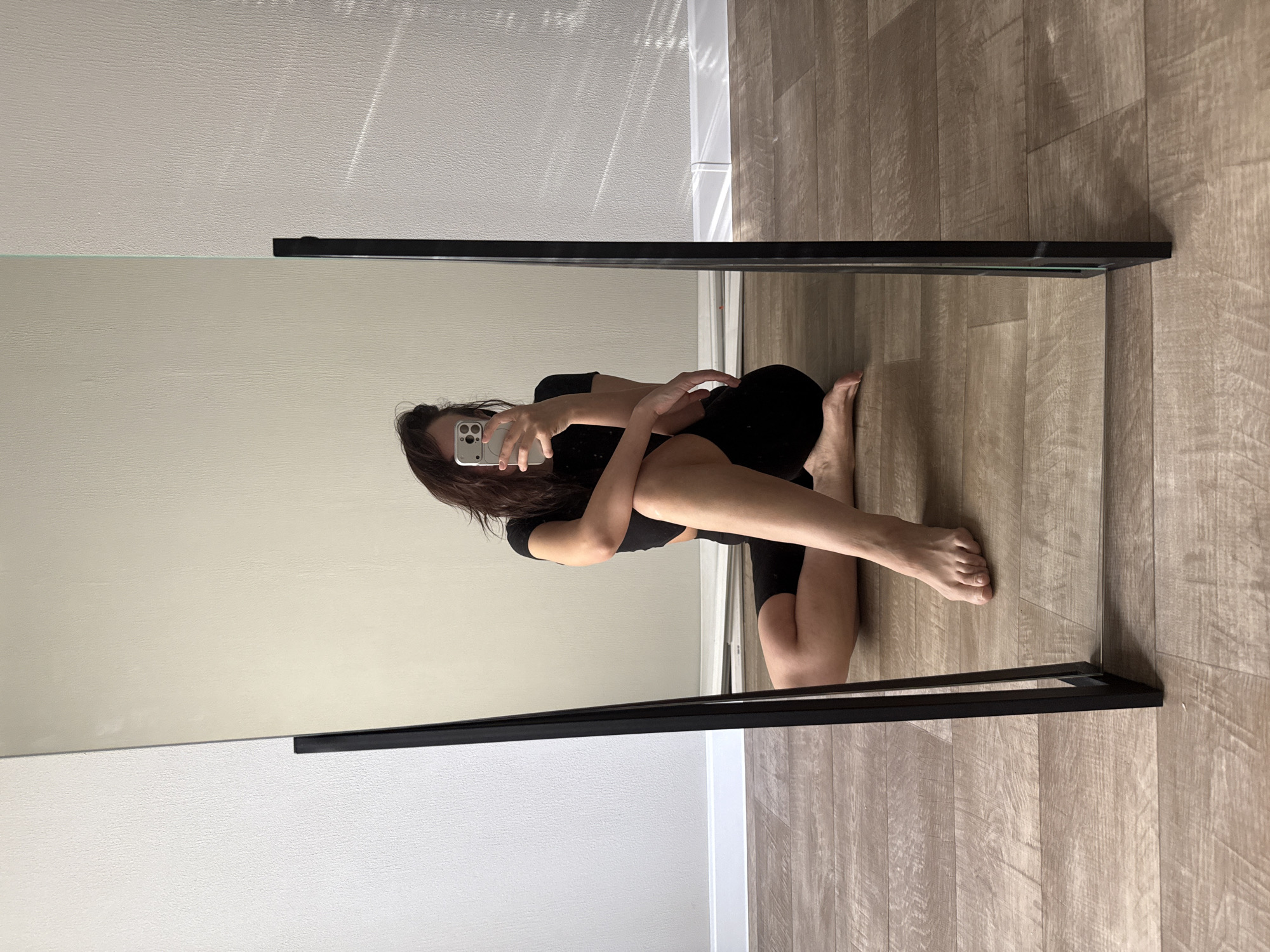The image shows a mirror selfie of a woman crouching on a wooden floor. She is wearing black athletic clothing. The background includes a white wall. The image does not contain information to conclude it was taken in Chaikovsky, Russia.