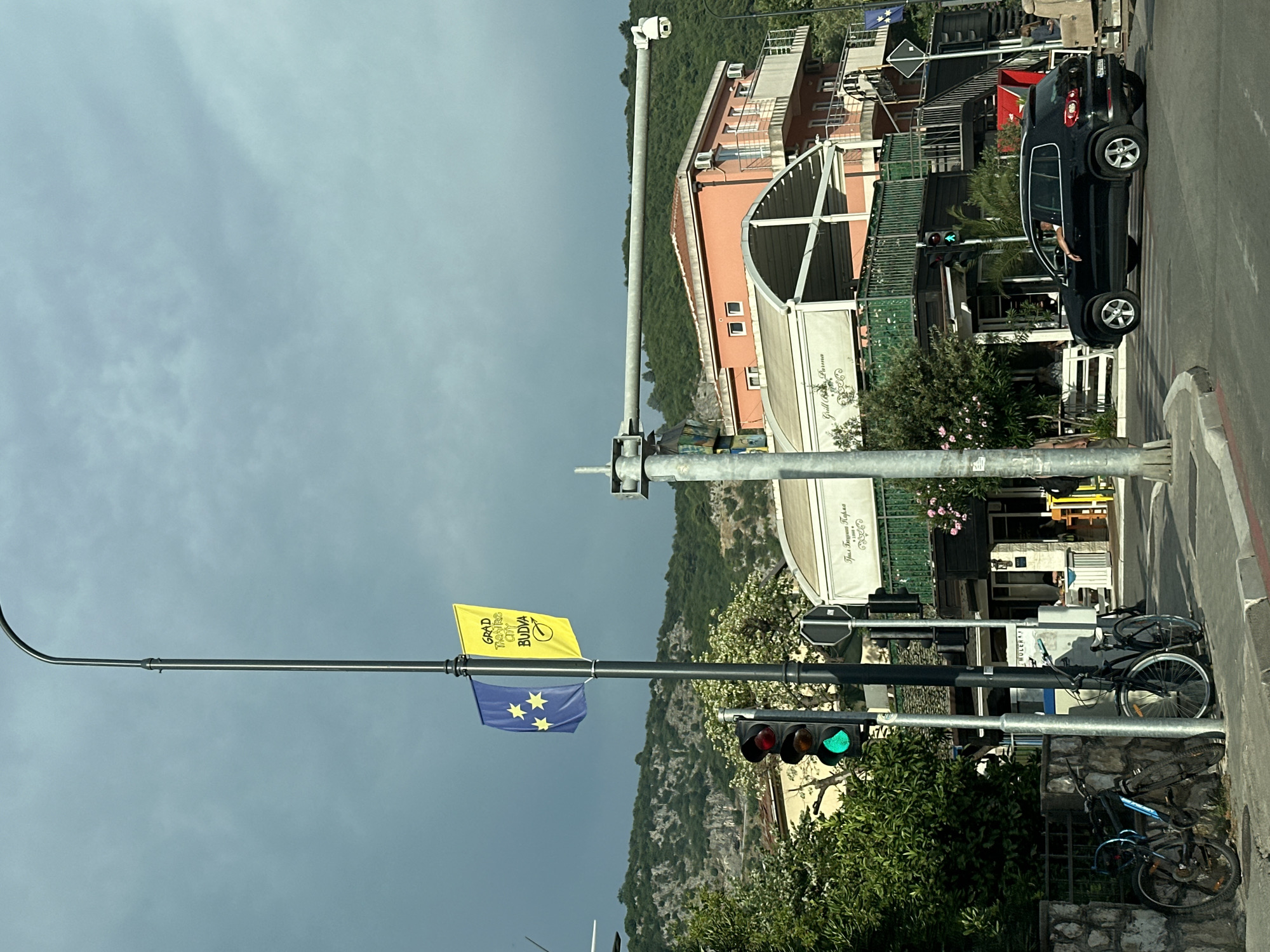 The image shows an outdoor scene in Budva, Montenegro. A street light with a traffic signal is prominent, showing a green light. Below the traffic signal, there are two flags hanging from poles. The top flag is yellow and displays text in what appears to be a Latin script, including "GRAD", "TRAFFIC", "CITY", and "BUDVA". The lower flag is blue with four yellow stars arranged in a square.
In the background, there is a black car parked on the right side of the road. Several buildings are visible, including one with a pink facade and another with a white awning that has some text on it. The scene is set against a backdrop of lush green hills and vegetation, suggesting a possibly sunny day despite the overcast sky. A few bicycles are parked near a stone wall. The overall impression is a street view in a coastal town. The time of day appears to be daytime.