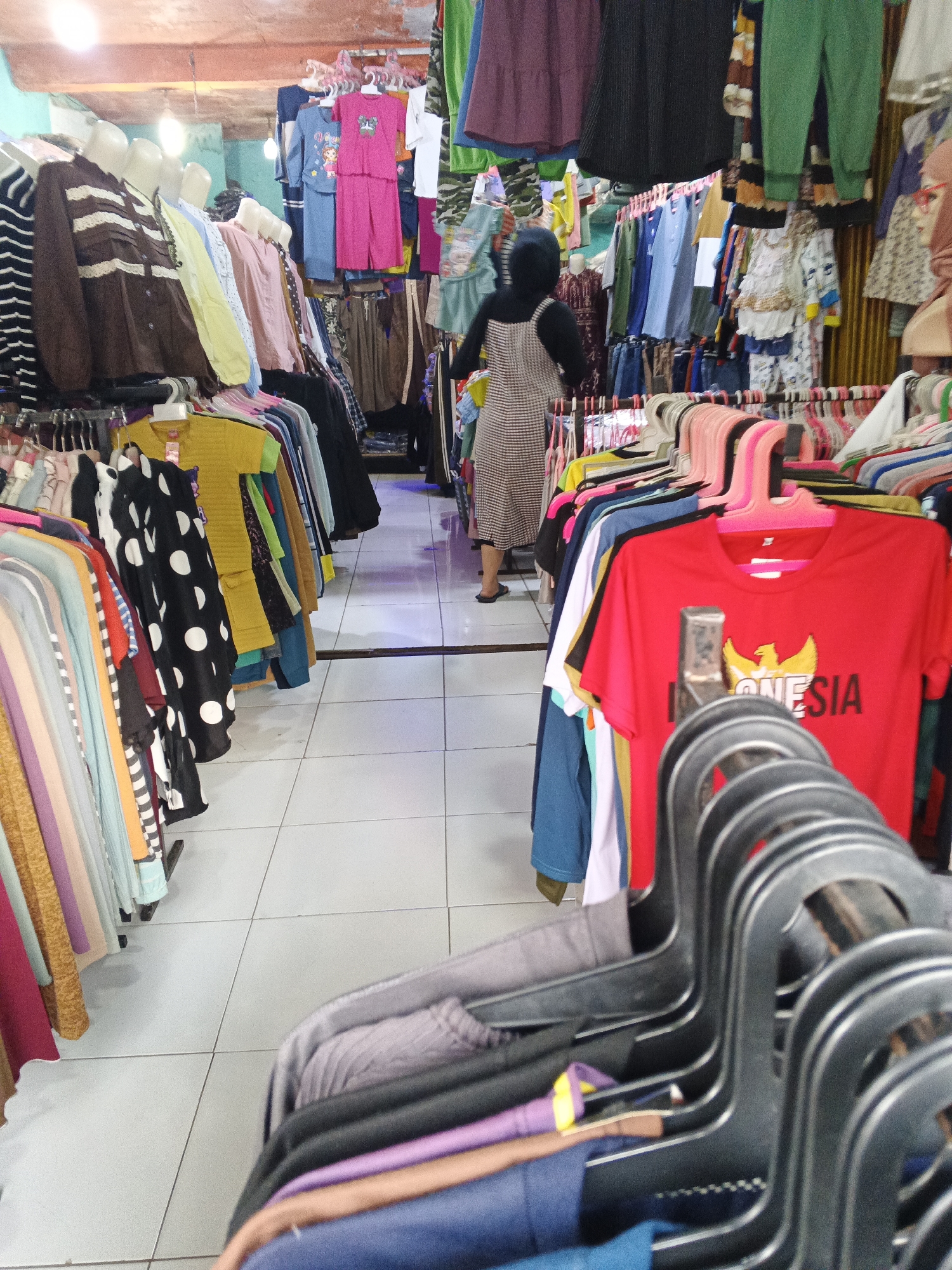 The image depicts the interior of a clothing store, likely in Warungkiara, Indonesia. The store is filled with racks of clothing, both on hangers and on display. In the background, a person wearing a hijab and a patterned dress is walking away from the camera. The clothing consists of various types, including shirts, pants, and dresses, in a range of colors and styles. The flooring appears to be tiled. The lighting suggests an indoor setting.