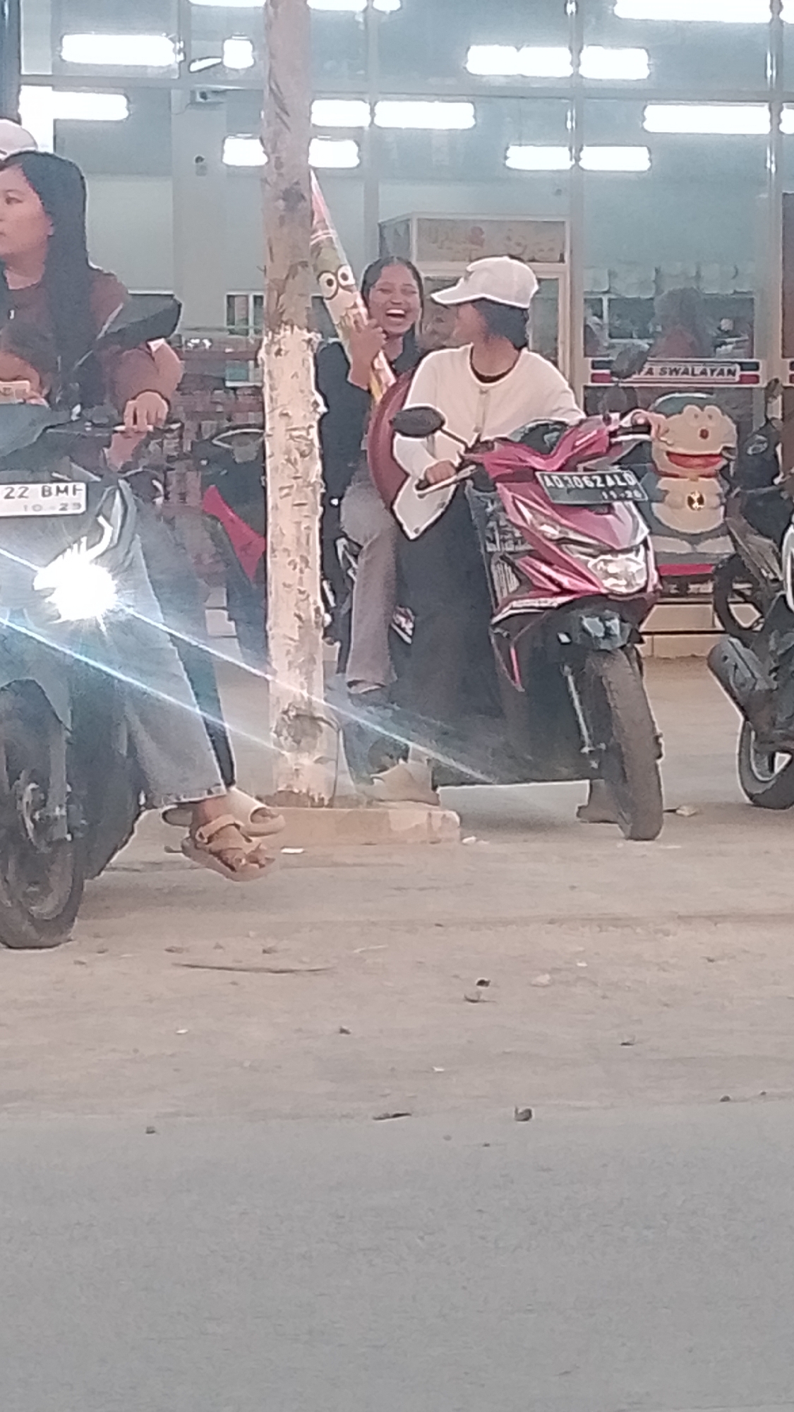 Two young women are on motorbikes in front of a building with a glass facade. The woman on the left, wearing a brown jacket, is looking to the left. The woman on the right, wearing a white cap and long-sleeved white shirt, is laughing and holding a cartoon character figure. Her motorbike is pink. A third person, partially obscured, is also on a motorbike to the right. The ground is paved, and there are several other motorbikes visible. The building in the background appears to be a commercial establishment, possibly a minimarket, with signs suggesting it is located in Karanglangu, Indonesia. The lighting suggests it is daytime.