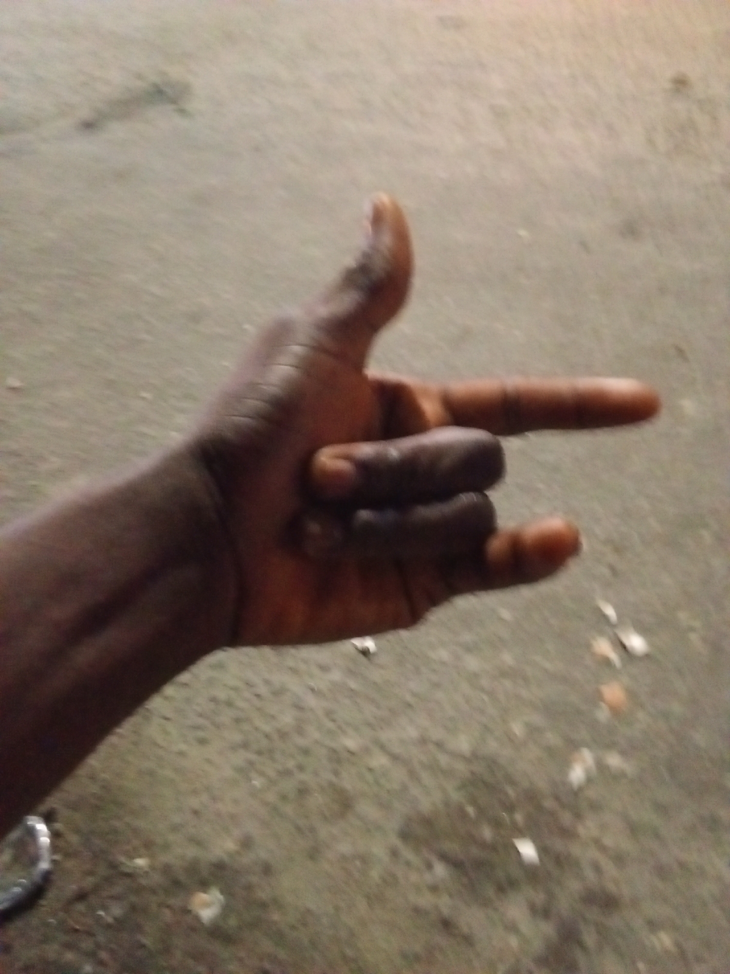 A person of African descent holds their hand in the shape of a gun, against the background of a concrete surface. The location appears to be Ado, Nigeria, based on image analysis indicating concrete surfaces typical of urban environments there. The background shows debris scattered on the concrete. There are no other people or objects visible. The primary action is the hand gesture. There are no interactions depicted. The environment appears to be an outdoor, urban area with concrete flooring.