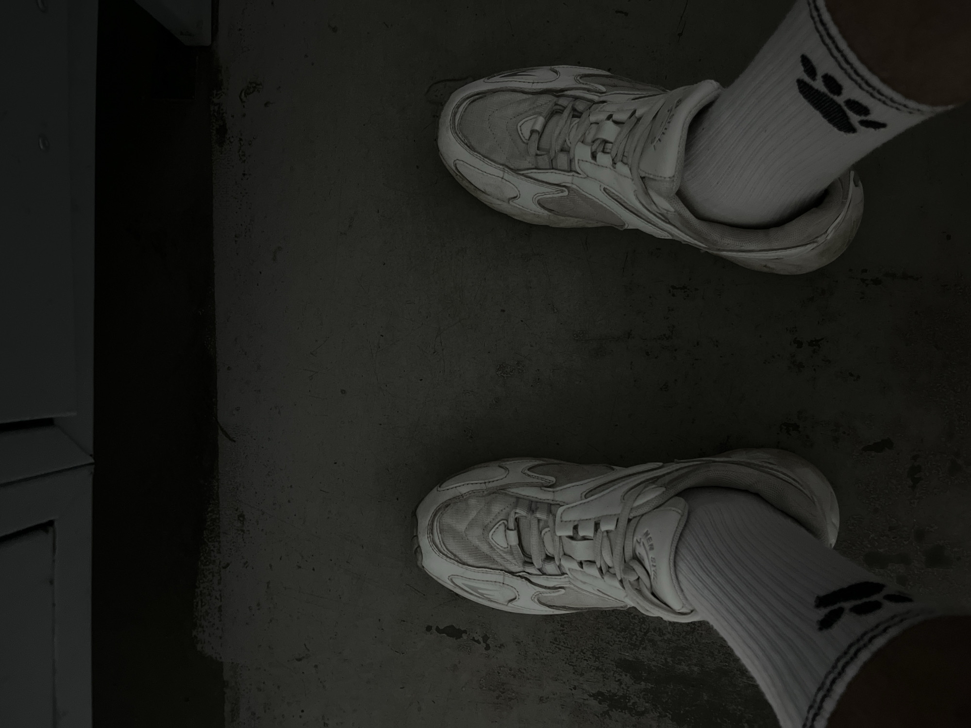 A pair of white athletic shoes with white socks featuring a black paw print design are shown from a top-down perspective. The shoes are laced and appear to be worn on a person's feet, standing on a dark, textured surface that resembles concrete or asphalt. The background is dark and out of focus, with a hint of a light-colored structure at the top. There are no specific indicators to place the location within Kyiv, Ukraine.