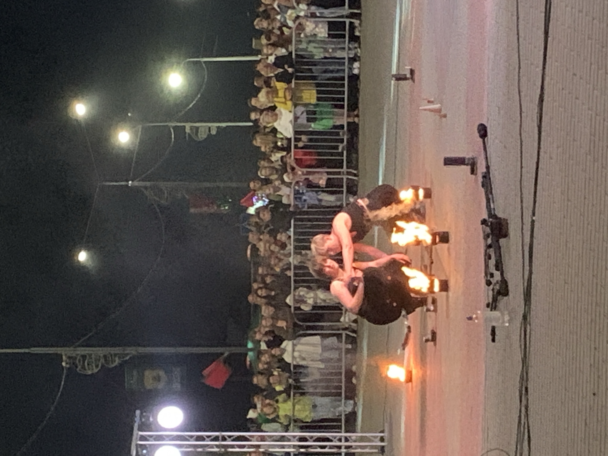 In Kapyĺ, Belarus, at night, a performance is underway with two individuals in dark attire kneeling amidst flames from torches. They are positioned in front of a large crowd of spectators, separated by a metal fence. The background features streetlights and stage lighting, indicating an outdoor event. Several small cones and cylindrical objects are placed on the ground around the performers, possibly part of the act. There's a bottle of liquid and some equipment in the foreground.
