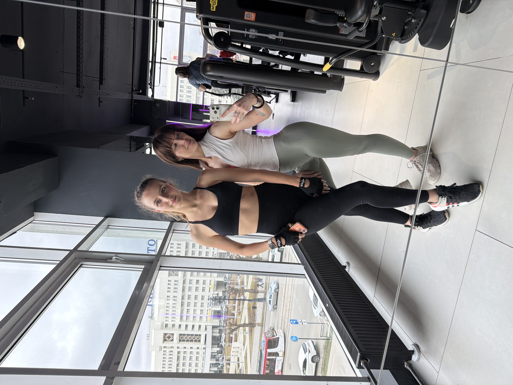 The image, oriented vertically, captures two women posing for a photograph inside a modern gym, likely situated in Perm, Russia. The setting features a dark industrial-style ceiling with exposed beams and track lighting, complementing a clean white tiled floor. Large floor-to-ceiling windows on the left side offer a view of an overcast sky and an urban street scene with multi-story buildings, vehicles, and a tram. One distant building displays a partially visible sign with the Cyrillic letters "ТО," while a vehicle on the street has "АВТОШКОЛА" (Avtoshkola - driving school) printed on its side.
The woman in the foreground, positioned slightly to the right, has blonde hair styled away from her face. She wears a black sports bra, black leggings, black athletic shoes with white soles and red accents (Adidas), and black gym gloves, holding a peach-colored smartphone in her left hand. Her expression is neutral as she looks towards the camera.
The second woman stands behind and slightly to the left, with brown hair and bangs. She is dressed in a white sleeveless top, light green ribbed leggings, and beige athletic shoes. She holds a silver smartphone in her right hand, making a "peace sign" gesture, and wears a black bracelet. Both women appear active and fit. Behind them, black gym equipment, including a machine with a "Technogym" logo and a "Scan me" QR code, is visible. The gym's interior also features purple accent lighting. The natural light from the windows suggests it is daytime.