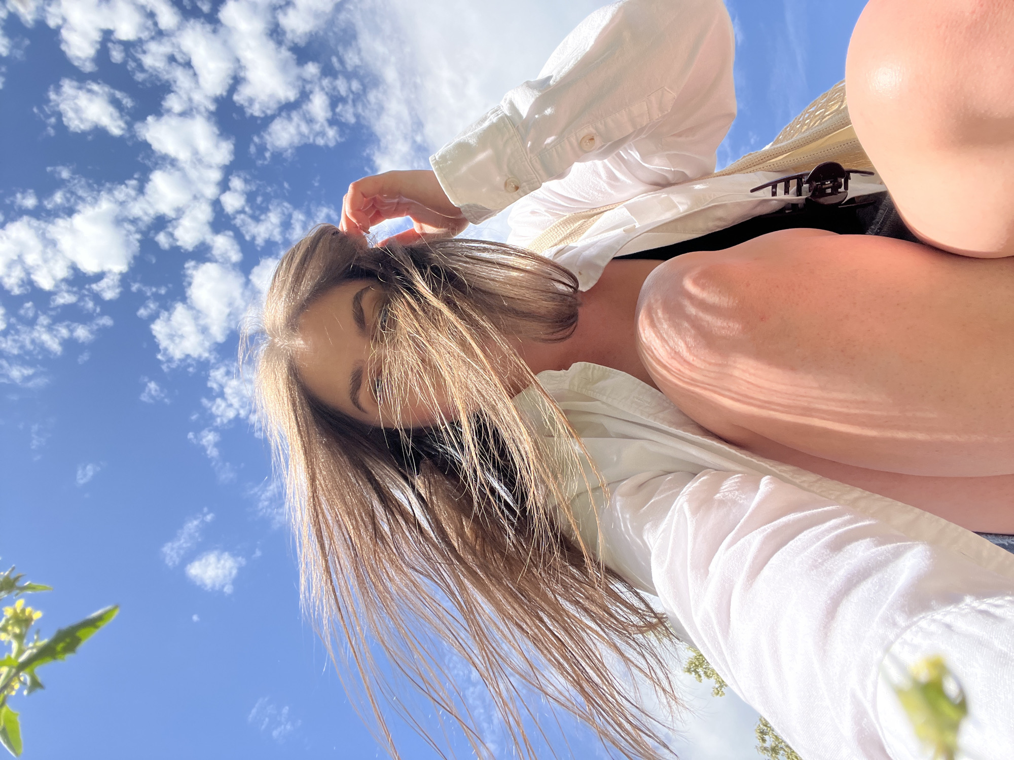 A low-angle, upward-facing shot captures a woman partially obscured by her own hair. She is wearing a white button-down shirt, and a black item of clothing is visible beneath. Her right hand is touching her hair. The background is a blue sky with scattered white clouds. Green foliage is visible at the bottom left corner. The location is unknown and can not be determined to be Ternopil, Ukraine from the given image.