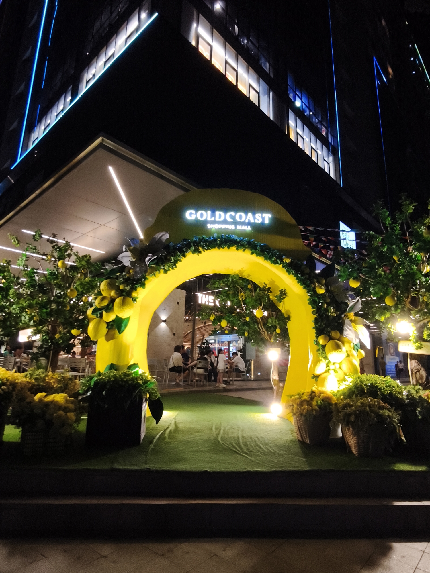 The image shows a nighttime scene outside a building with blue and white neon lights accentuating its modern architecture. In the foreground, a large, bright yellow archway decorated with artificial lemons and greenery frames an entrance. Artificial lemon trees flank the archway, laden with yellow fruits. The ground within the archway is covered in green artificial turf. Several people are seated at tables and chairs in the background, suggesting an outdoor dining or seating area associated with the building, possibly a shopping mall. The words "GOLDCOAST SHOPPING MALL" are visible on the archway. The location is identified as Nha Trang Ward, Vietnam.