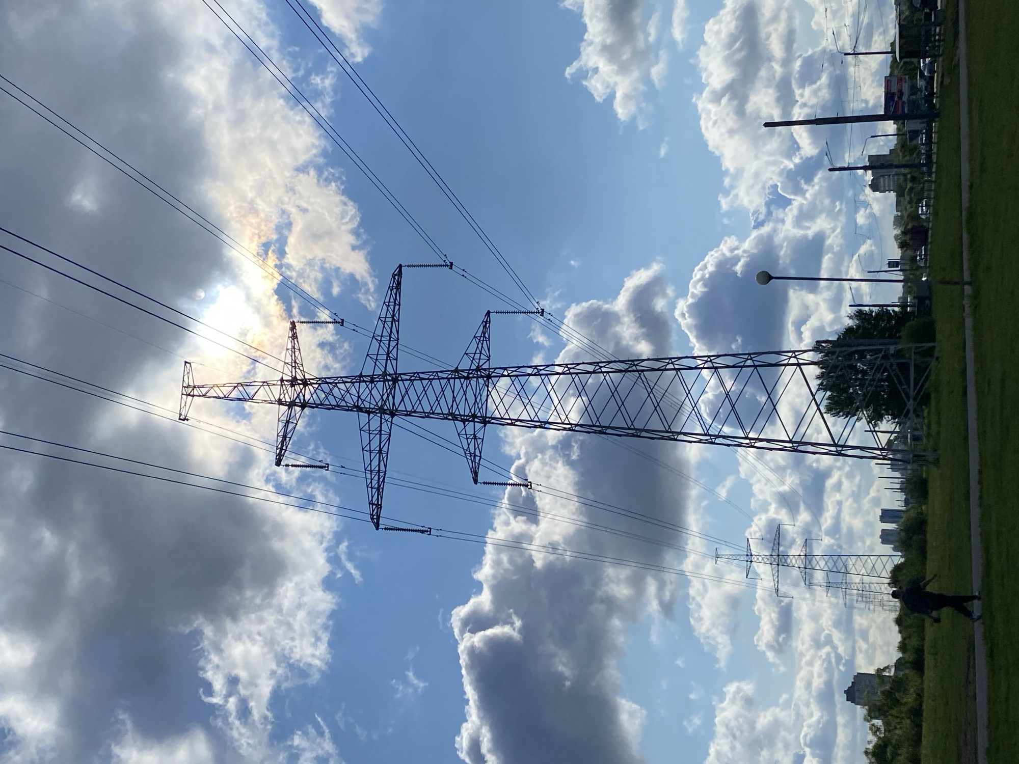 A wide-angle, low-angle shot captures a towering electricity pylon silhouetted against a dynamic sky filled with both bright sunshine and dramatic cumulonimbus clouds. The sun is partially obscured by the clouds, creating a lens flare effect and casting a warm, ethereal glow. Multiple power lines stretch diagonally across the frame, converging towards the pylon and disappearing into the distance. In the foreground, a person walks from left to right across a grassy area. Beyond the grassy field, a line of smaller pylons recedes into the background, suggesting a long power transmission route. A few buildings are visible on the horizon, indicating an urban or suburban setting. The overall impression is one of industrial infrastructure set against a dramatic natural backdrop.