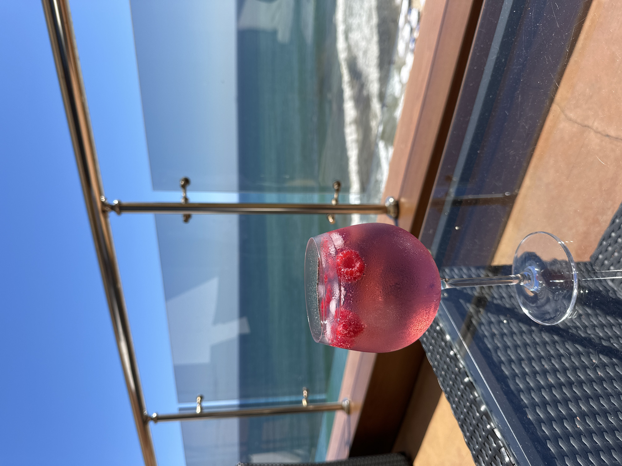 A glass of pink liquid with raspberries and ice sits on a glass-topped table on a balcony overlooking the sea. The glass is a wine glass with a condensation of water droplets on the outside. The liquid is a bright pink color and contains several whole raspberries and ice cubes. The table is made of woven black material with a glass top. In the background, the sea is visible under a clear blue sky. A railing with glass panels separates the balcony from the sea. The location is the city of Nesebar, Bulgaria, specifically on a balcony that offers a view of the Black Sea. The scene is a still shot, with no people or action depicted.