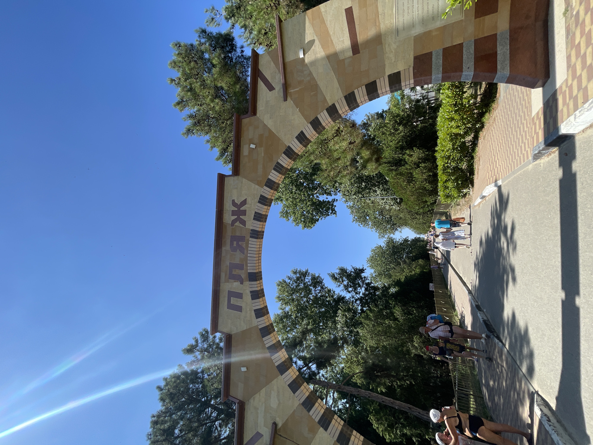 An outdoor, eye-level, wide shot captures a sunny day in Vityazevo, Russia, under a bright blue sky with lens flare effects from the sun. The foreground is dominated by a large, decorative archway with the word "ПЛЯЖ" (Beach in Russian) inscribed in Cyrillic letters across its top. The archway is constructed of stone blocks in varying shades of tan, brown, and cream, with a decorative pattern of dark brown and tan stripes on the arch itself.
Beyond the archway, a paved pathway leads into the distance, lined with lush green trees and bushes on either side. Several people are seen walking along the pathway, some heading towards the beach and others away from it. On the left side of the pathway, near the arch, a group of people is visible, including a woman in a black bikini and a white swim cap, and another woman with her arm around her. Further down the path, a man in a checkered shirt and shorts, carrying a blue bag, walks with a child.
In the background, more people are walking along the path, and the trees create a dense canopy, casting dappled shadows on the ground. A metal fence runs along the left side of the pathway. To the right of the archway, a stone monument with an inscription in Russian is partially visible. The overall scene suggests a warm, summer day at a beachside resort.