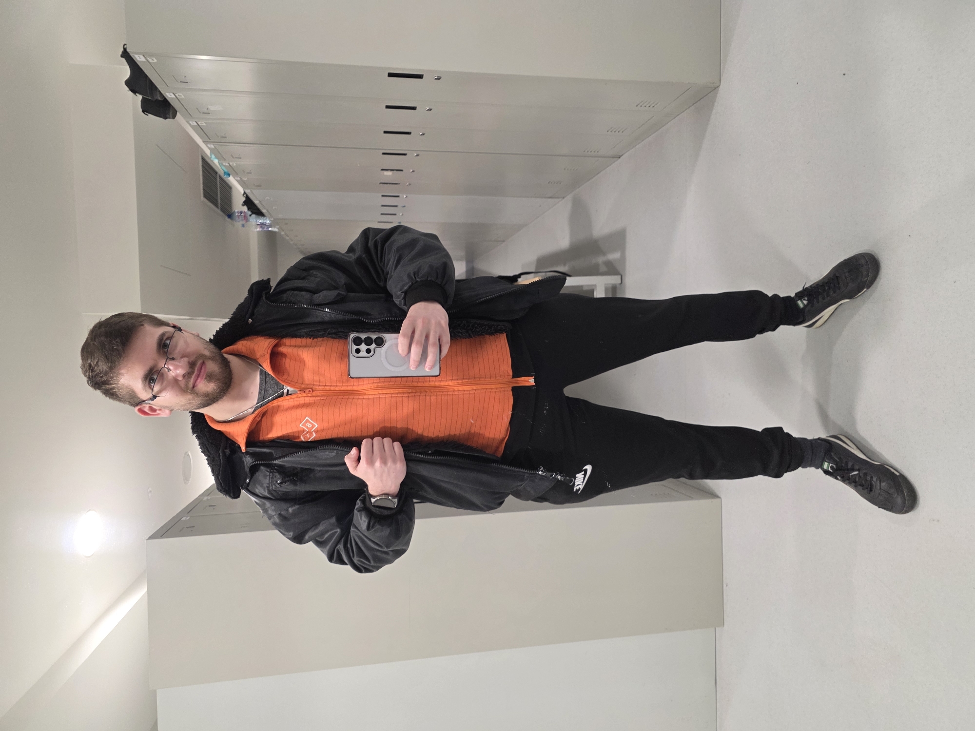 A man stands in what appears to be a locker room or storage area with rows of metal lockers stretching behind him. He is wearing glasses, an orange zippered jacket, black Nike sweatpants, and dark-colored shoes. He also sports a black jacket and a watch on his left wrist. He is holding a silver cell phone in his hands, possibly taking a selfie. The room has white walls and what seems to be a fluorescent light source overhead. The location, city of Plovdiv, Bulgaria is not identifiable from the image.