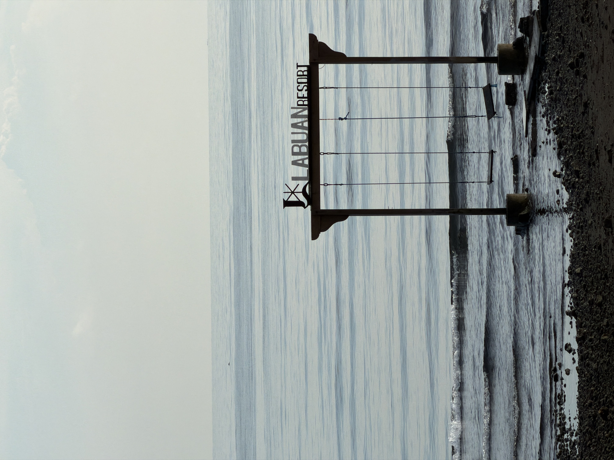 An outdoor, eye-level, medium shot captures a beach scene at the Labuan Resort in Pasir Putih, Indonesia. The image is dominated by the calm, blue ocean stretching to the horizon, with faint horizontal lines indicating gentle waves. A wooden swing set with two swings, partially submerged in the water, stands prominently in the foreground. Above the swings, a metallic sign reads "LABUAN RESORT." The shoreline is covered with dark pebbles and some water. The sky above is a pale blue with a few wispy clouds. The lighting suggests it is either early morning or late afternoon. There are no people visible in the image.