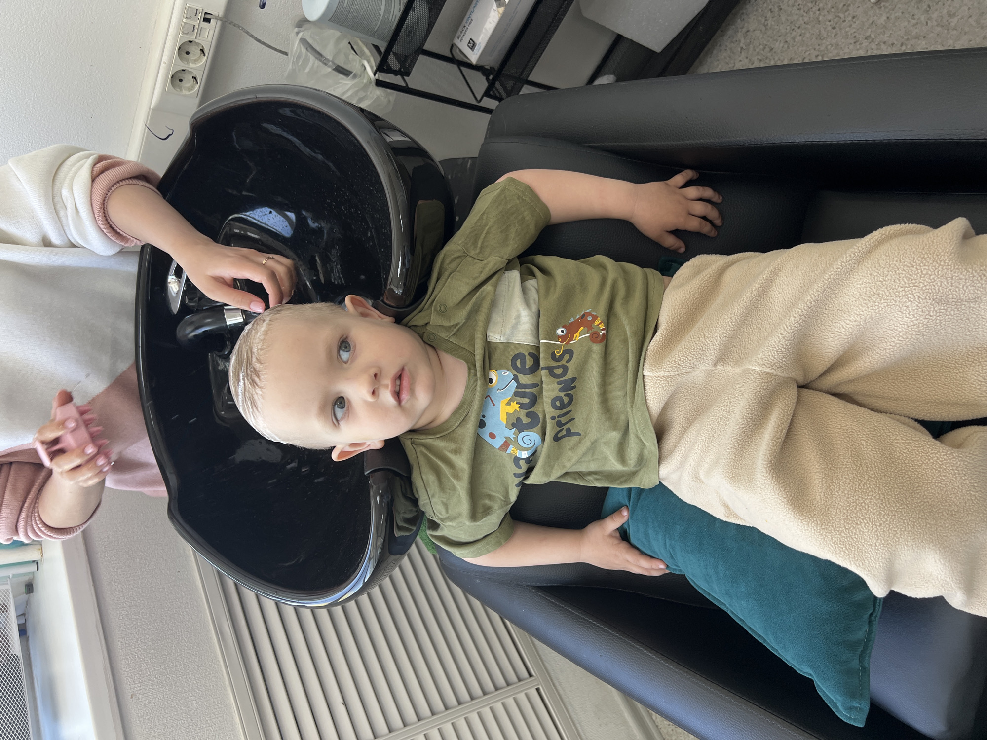 A young child is lying in a black salon chair getting their hair washed in a black basin. The child is wearing an olive green shirt with a "Nature Friends" design and beige pants. They are resting on a teal pillow. An adult is washing the child's hair. The setting appears to be a salon, possibly in Chaikovsky, Russia. There is a metal shelving unit in the background. The environment suggests a grooming process.