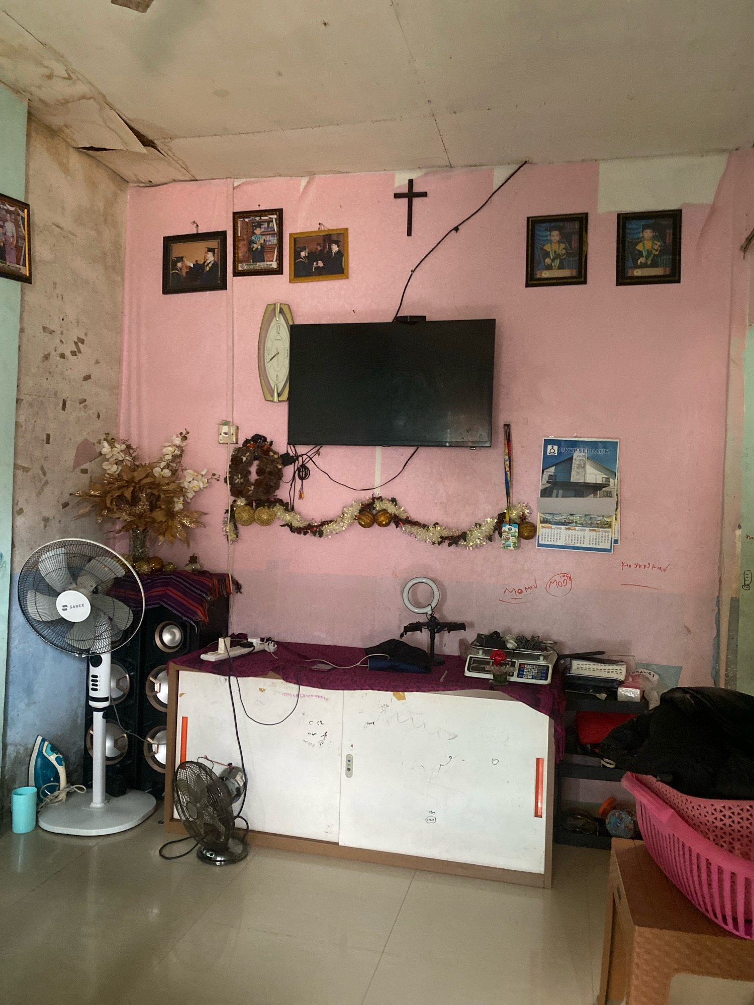 The image depicts an interior scene in a room that appears to be a living area or a multipurpose room, located in Batam, Indonesia. The room has pink walls and a damaged ceiling with visible peeling paint and water stains.
Dominating the scene is a large flat-screen television mounted on the pink wall. To the left of the television, a large standing fan with a white base and black blades is positioned on the floor. Next to the fan, there is a small turquoise cup and a blue iron. Behind the standing fan, a large black speaker with silver accents is visible.
On the pink wall above the television, several framed photographs are hung, some depicting individuals in graduation attire. A wooden cross is also mounted on the wall. Beneath the television, a decorative garland with white and gold elements is draped across the wall, along with a wreath. A wall calendar from 2024 is hanging to the right of the garland, featuring images of buildings.
In the center of the room, in front of the wall, is a white cabinet with drawers, topped with a dark purple cloth. On the cabinet, various items are placed, including a digital scale with a small red flower in a vase, a ring light on a stand, and other electronic devices and wires. A smaller, round fan is placed on the floor in front of the cabinet.
To the right of the cabinet, a dark shelf unit contains several items, including what appears to be a black trash bag and a pink plastic basket filled with items. The floor is tiled with light-colored tiles.
The overall impression is of a lived-in space with a mixture of functional and decorative items. The condition of the ceiling suggests potential issues with the building's maintenance.