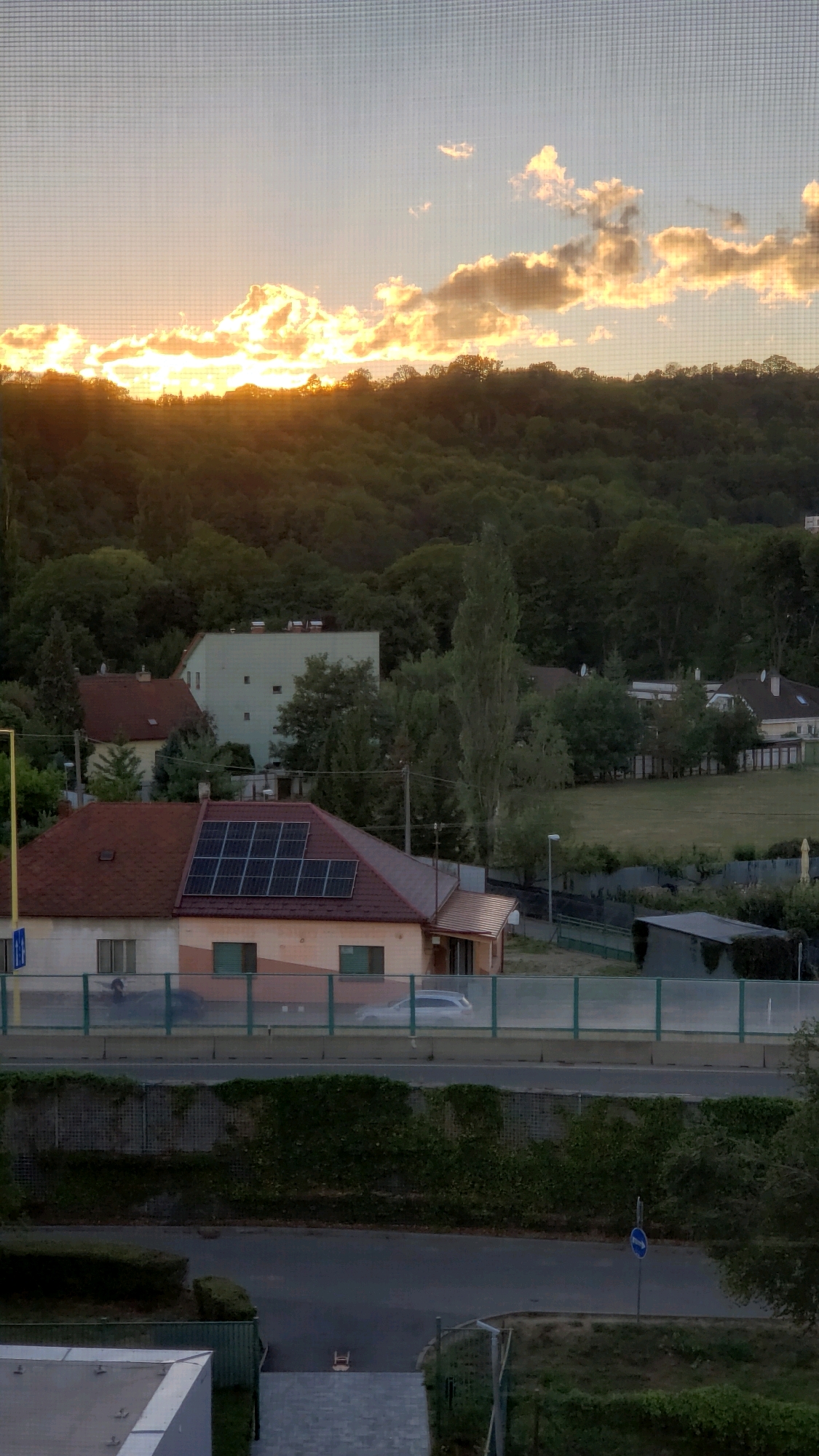 The image shows a residential area with a hill covered in trees in the background. In the foreground, there are several houses, one of which has solar panels on its roof. A road with a car driving on it is visible in the middle ground, separated from the houses by a barrier. The sky is partly cloudy with the sun setting behind the clouds, casting a warm glow over the scene. The overall impression is one of a peaceful suburban setting at dusk. The image captures a view of a street and houses in the city of Prešov, Slovakia.