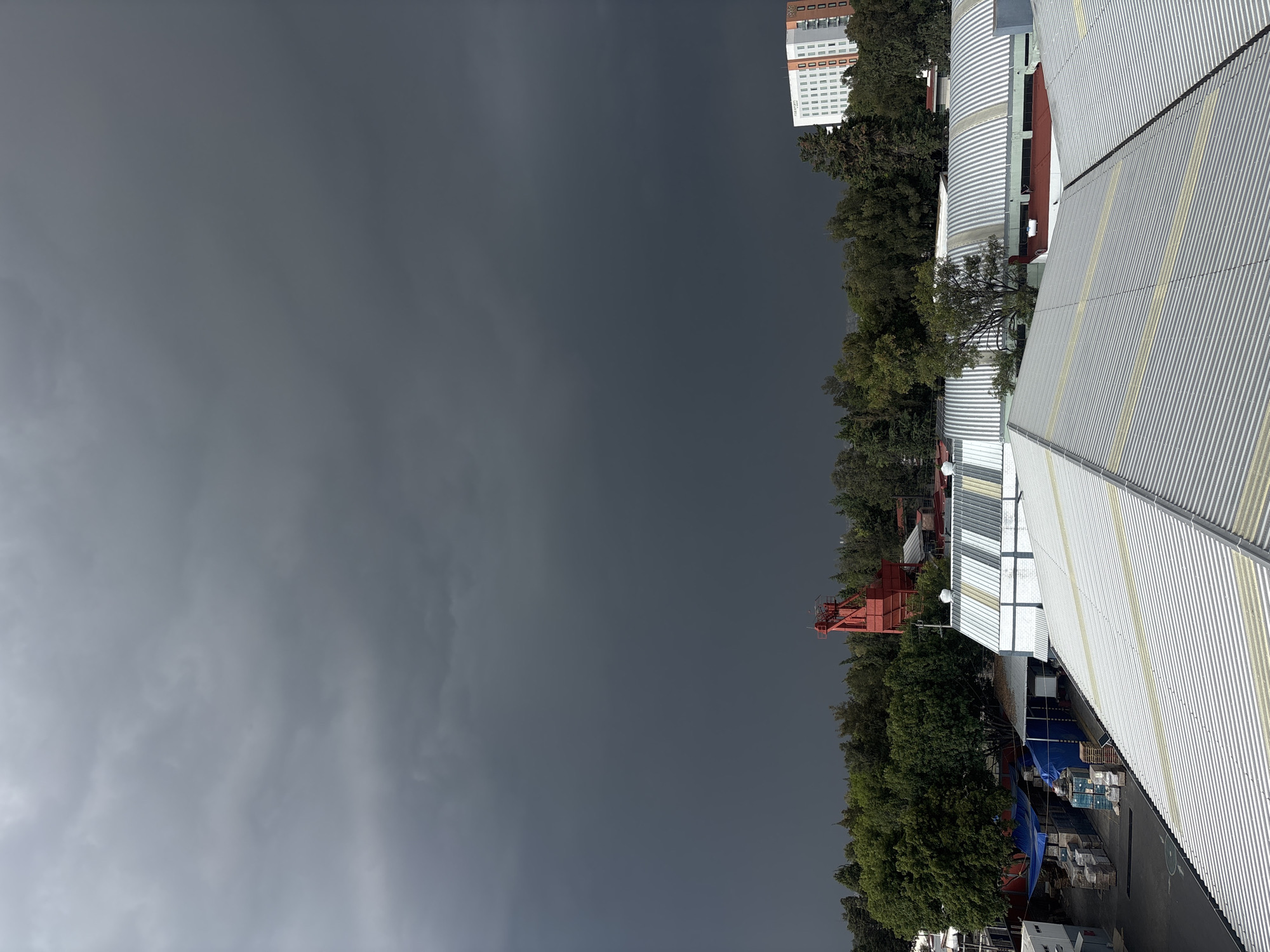 The image shows an overcast sky over a location in Puebla, Mexico. To the right of the frame are buildings with metal roofing, trees, and a tall white building in the background. A red industrial structure is visible among the trees. The dominant feature is the dark, cloudy sky.
