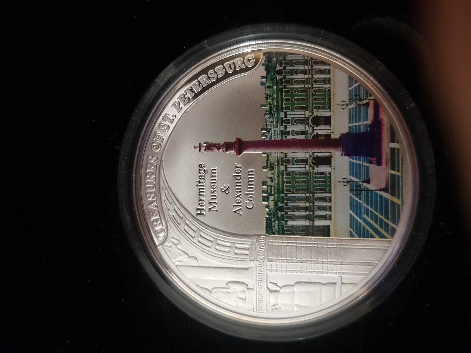 The image shows a commemorative coin featuring the city of Saint Petersburg, Russia. The coin's design highlights the Hermitage Museum and the Alexander Column. The words "Treasures of St. Petersburg" are inscribed around the coin's upper edge. The design incorporates elements depicting both the museum's architecture and the iconic column, combining them in a single image.