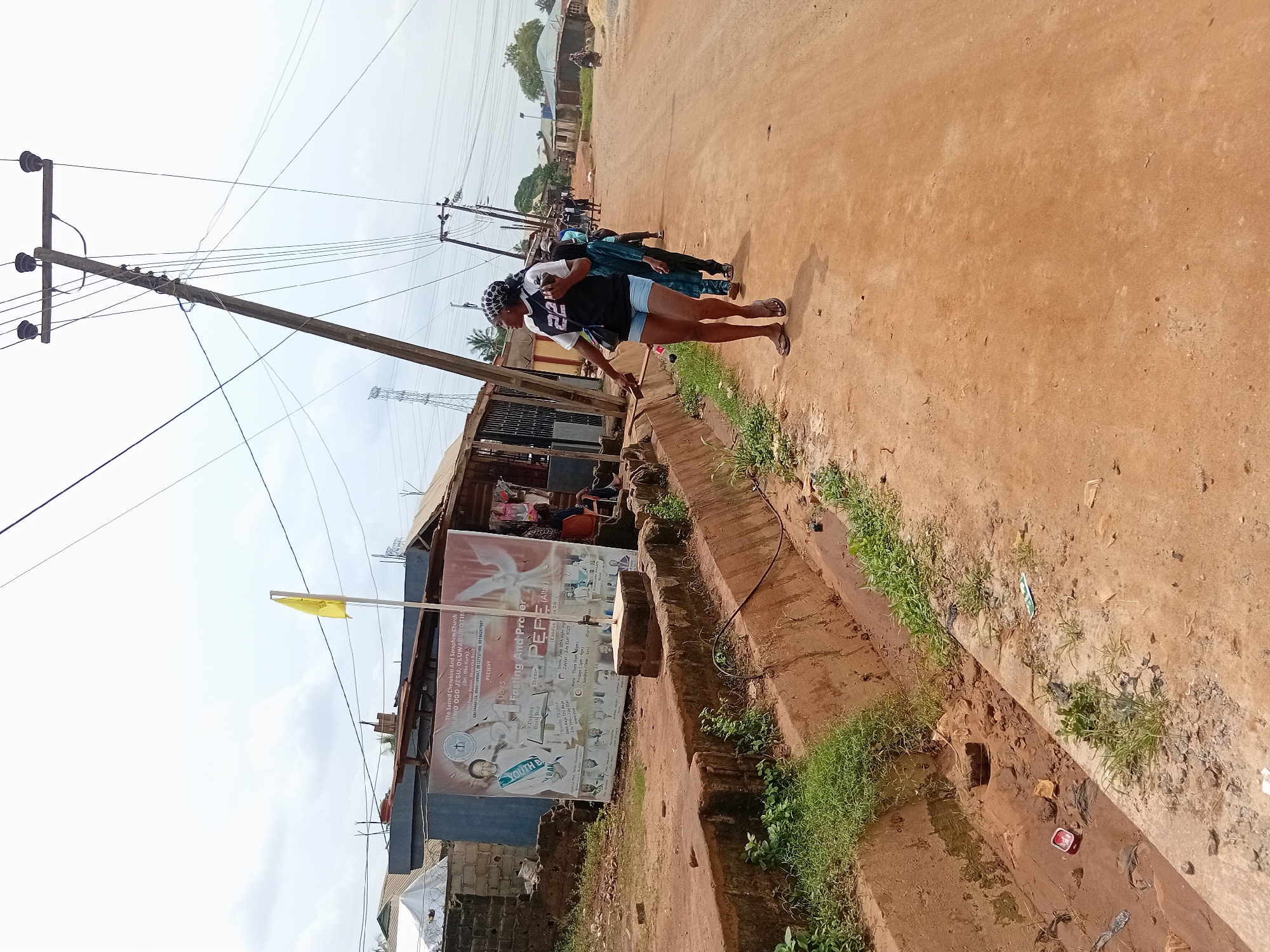 The image shows an outdoor scene in Ikorodu, Nigeria. The primary subjects are individuals walking on a dirt road or roadside. A woman, identifiable by her attire, is the most prominent figure. The environment includes utility poles with electrical wires, and a building displaying a sign advertising a religious event for "The Sacred Cherubim And Seraphim Church RAWO OGO JESU, OLUWADUROTIN," taking place from June 30th to July 21st, 2025. The scene appears to be a street with basic infrastructure. Vegetation is present along the roadside.