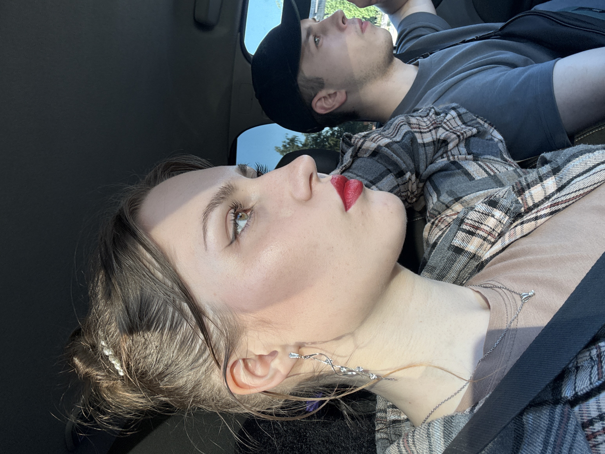 Two young adults are seated in a vehicle in Saint Petersburg, Russia, under direct sunlight.
The female in the front passenger seat is viewed in profile, facing towards the right side of the vehicle. She has brown hair styled in a messy bun, adorned with a small clip featuring multiple light-colored beads. Her visible left ear has a long, silver-toned earring with a rectangular stud and a twisted design inlaid with clear stones. A thin silver chain necklace with a single-stone pendant rests on her neck. Her eyes are light-colored, and she wears black winged eyeliner and red lipstick. She is dressed in a light-colored t-shirt beneath an open grey, white, and brown plaid flannel shirt, secured by a black seatbelt across her chest.
To her right and slightly behind her, the male driver is also in profile, facing forward. He wears a dark grey t-shirt, a black baseball cap worn backward, and a black shoulder bag across his torso. His right arm is partially visible on the steering wheel.
The interior of the vehicle is dark. Through the car windows, a blue sky and sparse green foliage are visible, indicating an outdoor setting, likely urban or suburban. Strong sunlight illuminates the female's face and shoulder, creating distinct highlights and shadows.