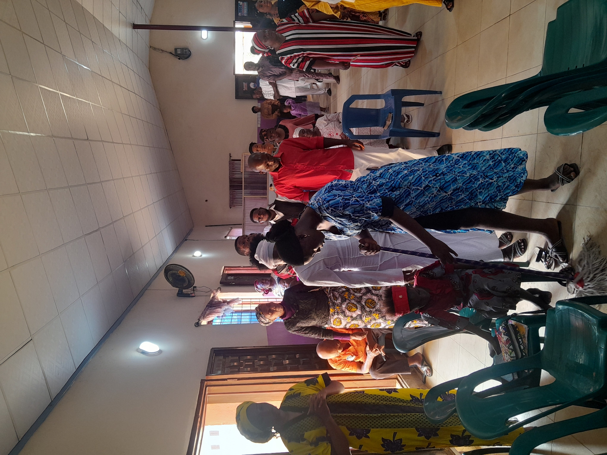 In a well-lit interior, possibly a church or community hall in Jos, Nigeria, a group of approximately 15 people are gathered. Most of them are adults, and they are dressed in colorful, traditional African attire. Several are wearing dresses or garments with vibrant patterns and designs, while others are in solid-colored shirts or wrappers. The people are standing or kneeling in a row, many of them appearing to be engaged in prayer or a religious activity.
The room features a tiled ceiling with recessed lighting, along with a wall-mounted fan. There are windows along one side, allowing natural light to filter into the space. Several blue and green plastic chairs are stacked or placed around the room. A broom leaning against one of the chairs near the center of the frame. A door is visible at the lower left corner of the image.
