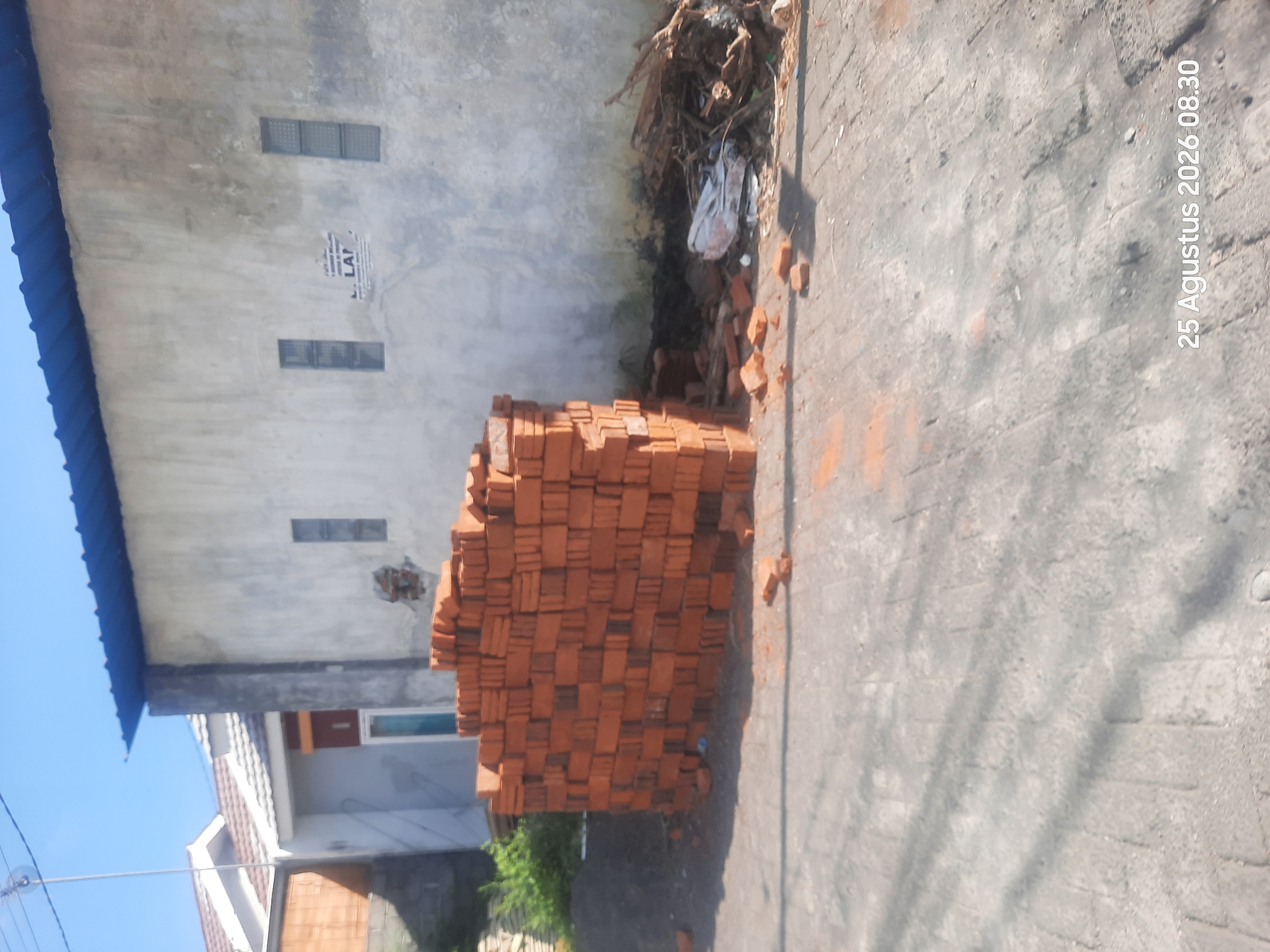 In Sukokerto, Indonesia, on August 25, 2026, at 08:30, an outdoor scene features a pile of red bricks stacked in front of a light gray wall of a building. The wall has a blue roof extending above it. There are two small, rectangular windows visible on the wall, one on the left and one on the right. To the left of the brick pile, some green foliage is visible, and further left, a portion of a residential structure with a white roof is present. To the right of the brick pile, there is a pile of debris consisting of wood and other materials. The ground is paved with interlocking blocks, and shadows are cast across it, indicating sunlight. No individuals are depicted in the image.