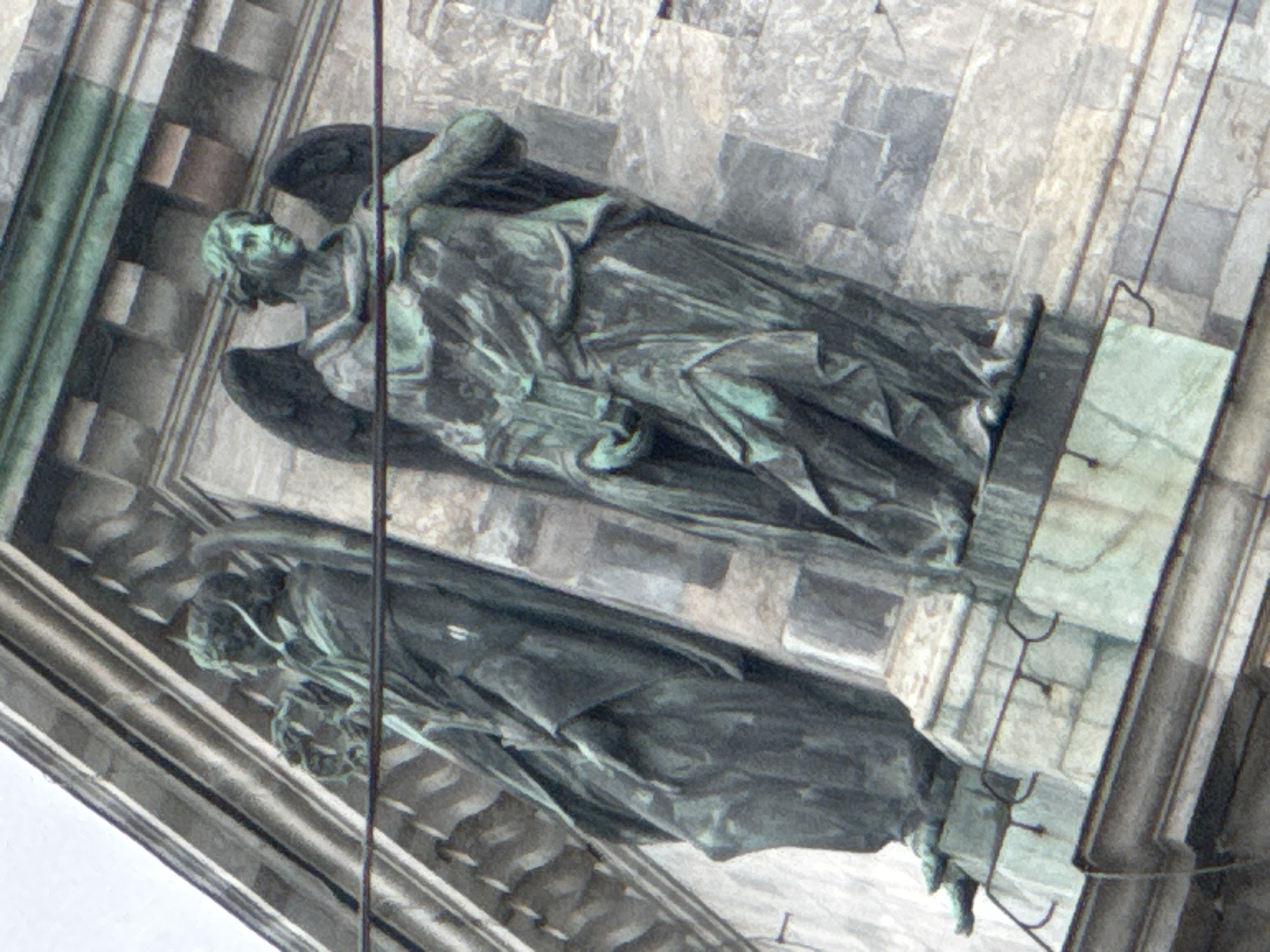 This image captures two large, weathered statues, likely made of bronze due to their distinct green-grey patina, set against a classical-style architectural facade in Saint Petersburg, Russia. The original image is rotated, making the figures appear to stand vertically, though they are likely positioned horizontally on a roofline, pediment, or a high architectural element of a grand building.
The upper statue is a winged figure, possibly an angel or an allegorical representation, depicted with finely sculpted hair, a serene expression, and elaborate, flowing drapery. It appears to be holding an elongated object, perhaps a scroll or staff, against its body. The lower statue is partially obscured but also shows a robed, draped figure, seemingly in a reclining or recumbent pose.
The statues are integrated into an ornate stone structure, featuring detailed masonry, columns, and decorative elements, characteristic of historical Saint Petersburg architecture, such as a cathedral or a palace. The weathered condition of both the sculptures and the building materials indicates significant age and prolonged exposure to the elements. The figures are static, representing monumental art rather than depicting an ongoing activity. There is no visible text in the image.