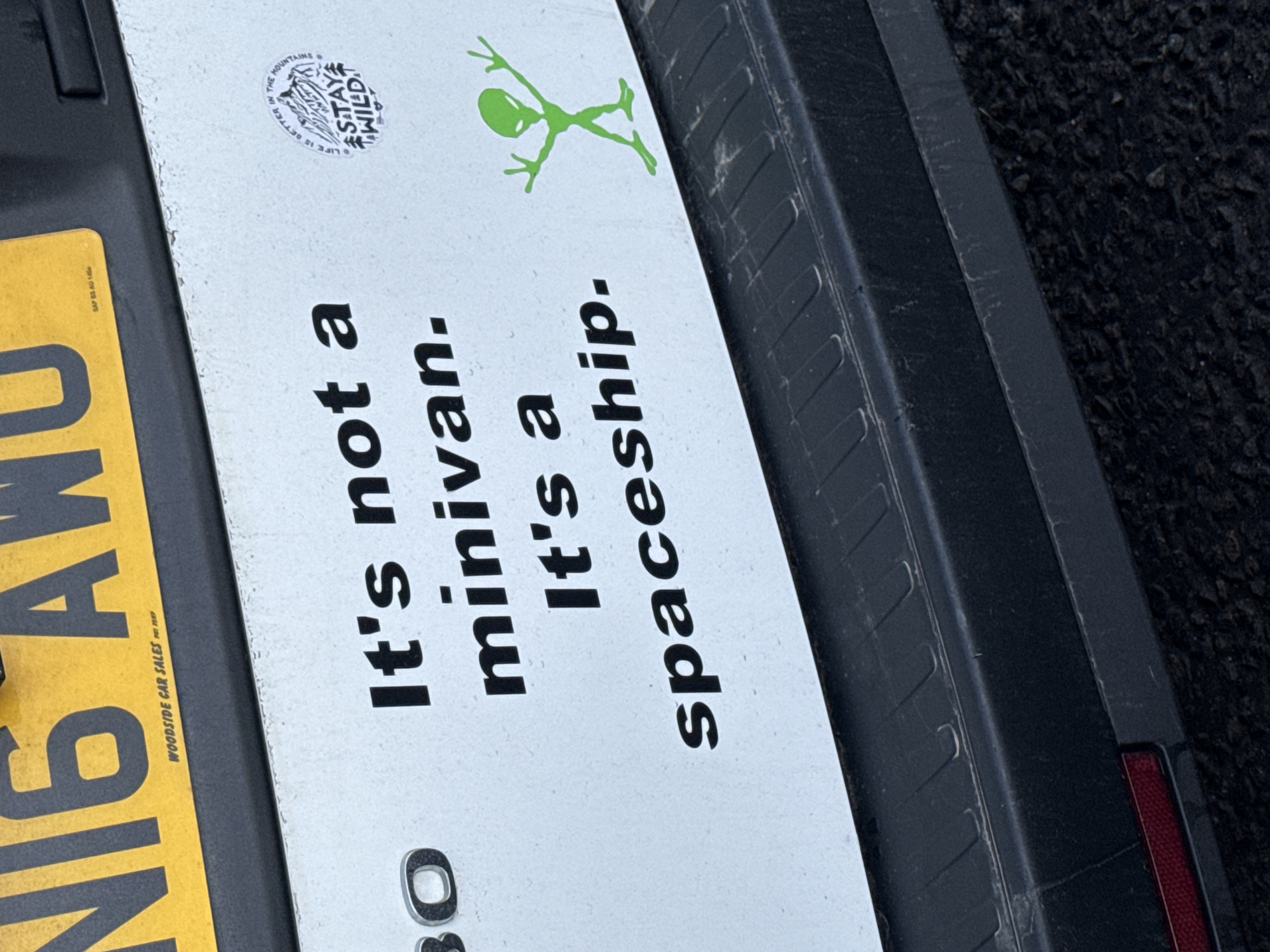 A close-up shot features the rear of a vehicle. On the vehicle's body, the words "It's not a minivan. It's a spaceship." are printed in black block letters. A green alien figure is situated above this text, adjacent to a sticker that reads "LIFE IS BETTER IN THE MOUNTAINS STAY WILD." The left side of the frame includes a partially visible yellow license plate with black text. The name "WOODSIDE CAR SALES" is imprinted on the license plate. The vehicle's rear bumper is also visible, showing a dark gray color and signs of wear and tear. The background is a dark, indeterminate surface.
