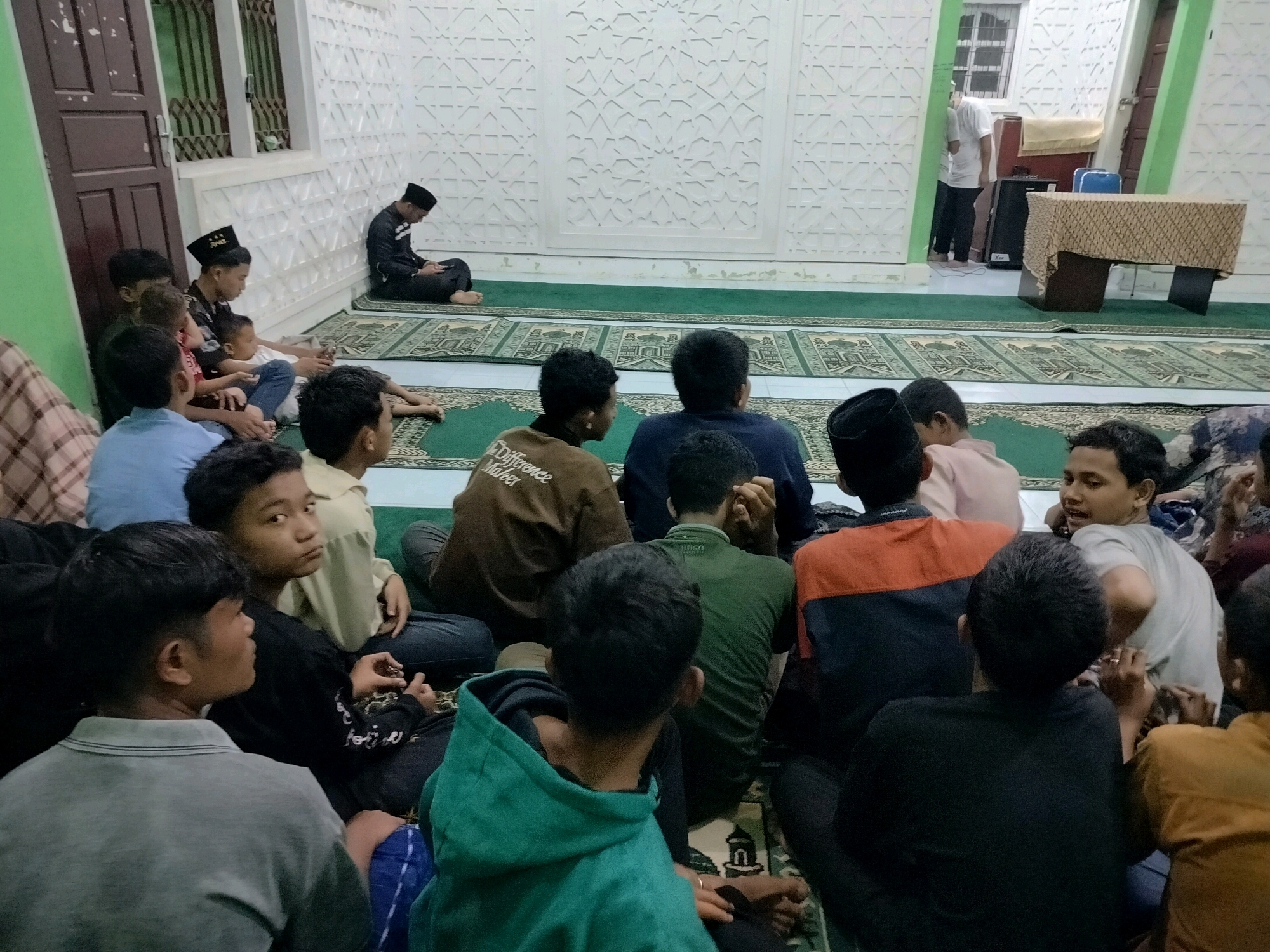 The image depicts an indoor gathering, likely in a prayer hall or community center in Lawebekung, Indonesia. Several young males are seated on green prayer mats arranged on a carpeted floor. In the background, a person in a black cap and robe is seated facing away from the camera, possibly engaged in prayer or reflection. Another individual, wearing a white t-shirt and dark pants, stands near a table covered with a patterned cloth. The walls are adorned with intricate white geometric patterns. The lighting suggests an ambient interior setting. The subjects are casually dressed, and the overall atmosphere appears peaceful and communal.