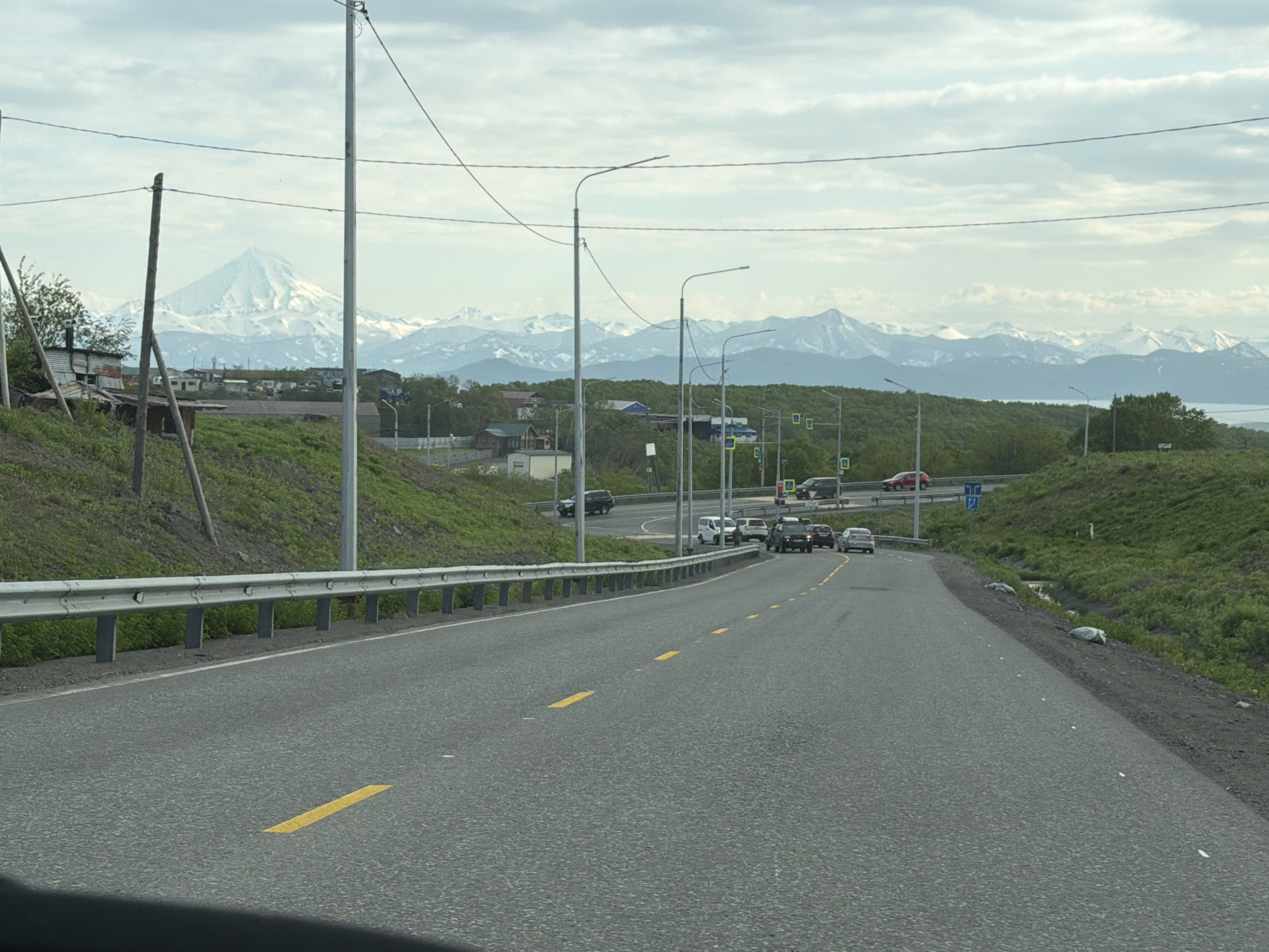 The image depicts a scenic view of a road in Krutoberegovy, Russia, leading towards a majestic mountain range capped with snow. The road, marked with yellow lines, curves gently to the right, with a metal guardrail on the left side. Several vehicles, including cars and SUVs, are driving on the road, suggesting a moderate level of traffic. The surrounding landscape is characterized by lush green hills and scattered trees, with a few buildings visible in the distance. The sky is overcast with clouds, indicating a possibly cool or temperate day. In the background, the imposing, snow-covered mountains create a dramatic backdrop, with a hint of the sea or a large body of water visible on the far right. The overall scene conveys a sense of natural beauty and rural tranquility, with hints of human presence through the road and vehicles. The time of day appears to be daytime, given the natural light.