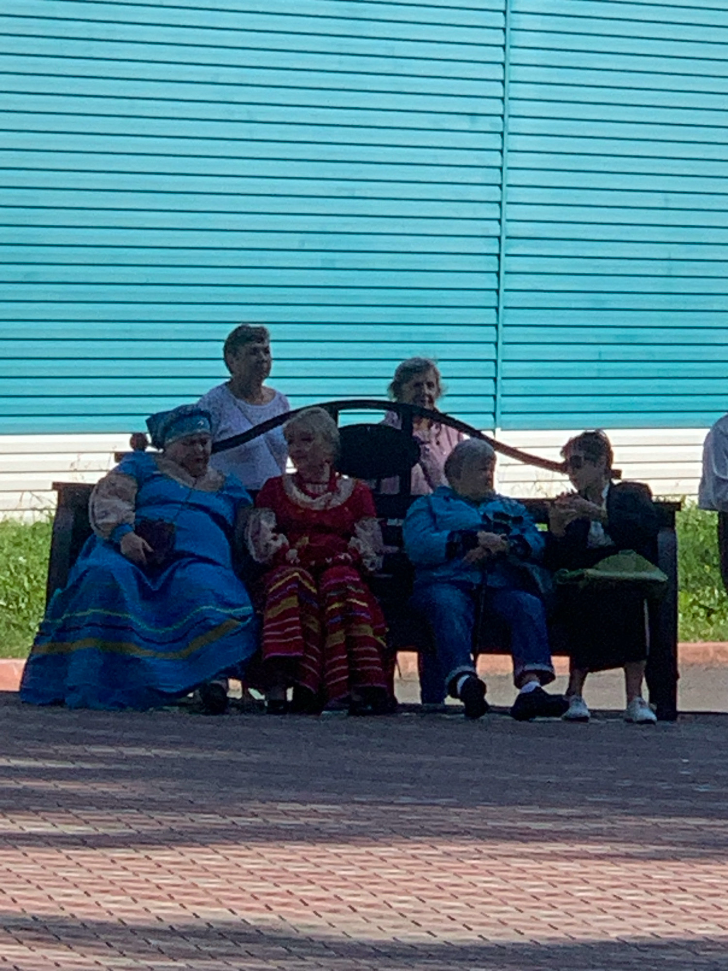 In Prokopyevsk, Russia, six elderly women are seated on a park bench in front of a bright teal, horizontal-slatted fence. The women are dressed in a variety of outfits. The woman on the far left wears a flowing blue dress with a matching headscarf. Next to her, a woman in a red and yellow striped dress with a white lace collar is seated. To her right, a woman in a blue jacket and pants is sitting, holding a cane. The fourth woman from the left wears a black jacket and skirt, and is looking down. The two women in the back are less visible but appear to be wearing casual tops. The bench appears to be dark wood and is positioned on a red brick pathway, with green grass visible behind the fence. The overall scene suggests a public gathering or outing for the women.