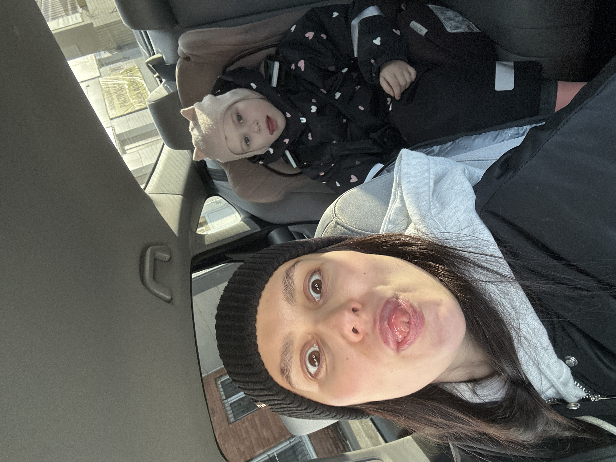 The image, rotated 90 degrees counter-clockwise, depicts a selfie taken inside a car in Chaikovsky, Russia.
**Subjects:** The foreground features a woman with dark hair and eyes, wearing a black ribbed beanie, a grey hooded sweatshirt, and a black jacket. She is captured mid-expression, sticking her tongue out playfully. In the backseat, a young child is reclined in a car seat. The child wears a light pink hat with ear-like details and a dark-colored jumpsuit adorned with numerous white and pink heart-like patterns. The child has fair hair and appears to look towards the camera with a calm, curious expression.
**Objects:** Visible car interior elements include grey fabric car seats, the child's beige car seat, and the car's grey headliner with a small grab handle.
**Scene and Setting:** The scene takes place within a vehicle, with bright natural light illuminating the subjects, indicating daytime. Buildings are visible through the car windows in the background.
**Activity or Event:** The woman is taking a selfie, capturing herself and the child in what appears to be a casual, personal moment during a car ride. Her playful facial expression contrasts with the child's more serene demeanor.
**Notable Details:**
* **Time of Day:** The strong sunlight suggests it is daytime.
* **Weather:** The bright, clear lighting implies sunny weather.
* **Location Cues:** Through the windows, an urban or residential environment with buildings can be seen.
* **Visible Text:** On a yellow sign or banner outside the car, partially obscured text includes "200" and what appears to be the Russian phrase "ХОЛОДНЫЕ НАПИТКИ" (meaning "COLD DRINKS").