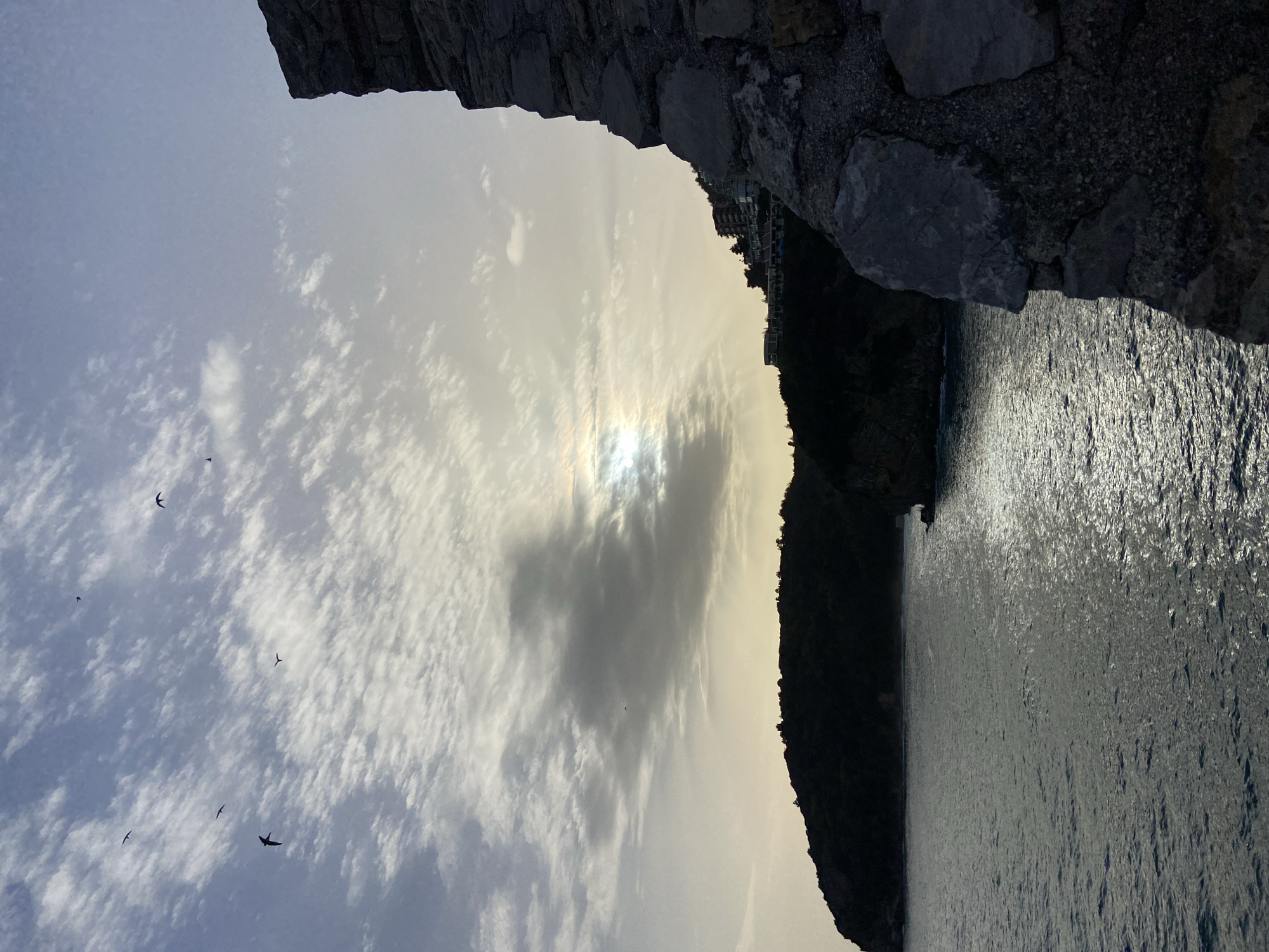 The image shows a coastal scene in Budva, Montenegro. In the foreground, a rough stone wall partially frames the view. Beyond the wall, a body of water, likely the Adriatic Sea, stretches towards the horizon with visible surface reflections. On the right side of the image, a dark, steep coastline is visible, with some buildings perched on its upper edge. Above, the sky is filled with a mix of patchy clouds and bright sunlight, with several birds flying across the sky. The sun is partially obscured by a dark cloud.