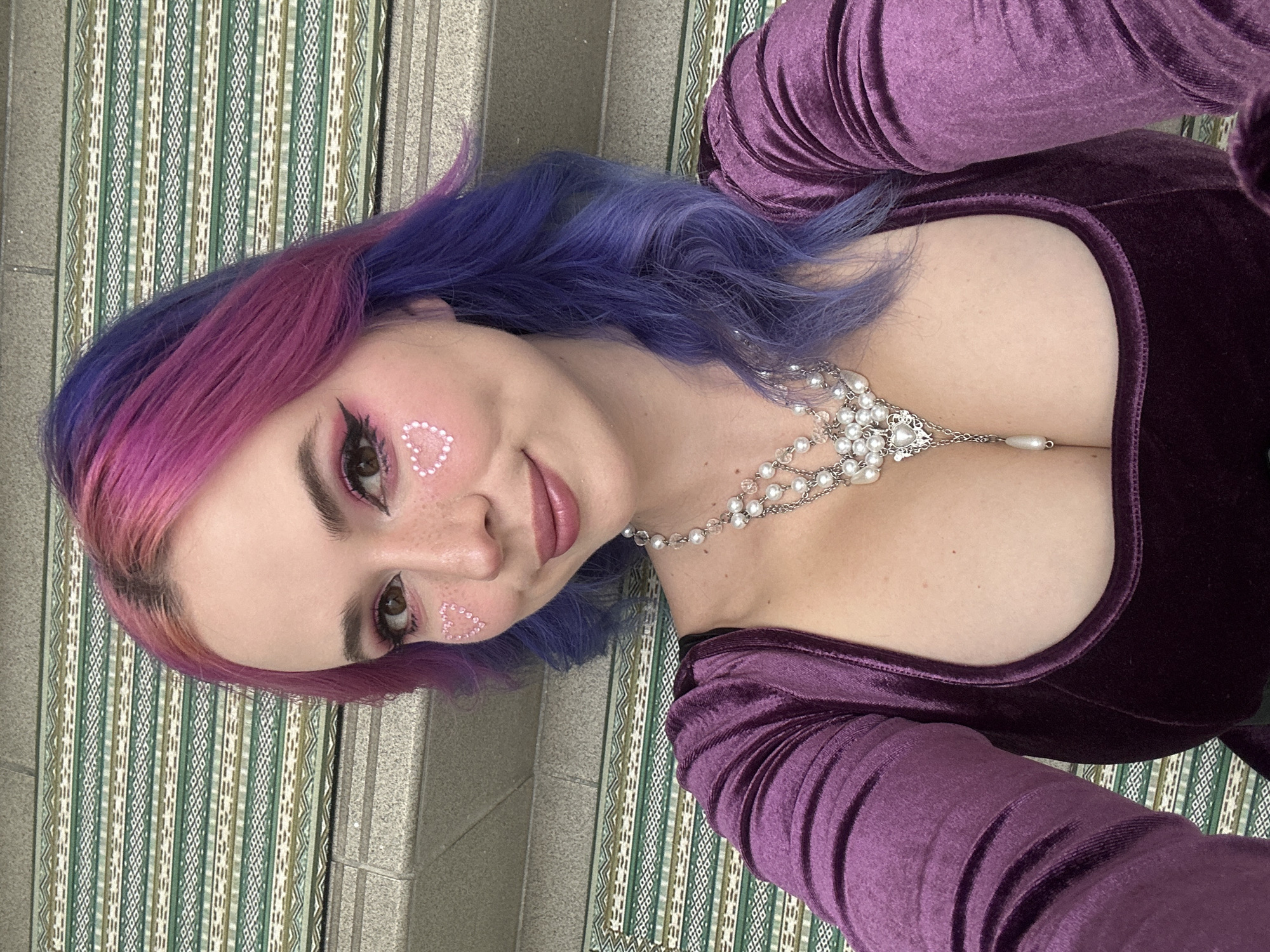 A woman with pink and blue hair is wearing a purple velvet dress and a pearl necklace. She has white heart-shaped designs on her cheeks. She is lying on a green and white striped fabric that is on a concrete surface, possibly a staircase. The location is unknown and there is no indication that it is in the city of Zagreb, Croatia.