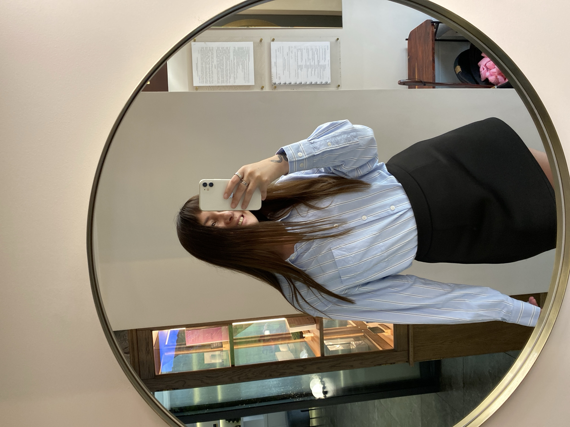A young woman is taking a mirror selfie in what appears to be a retail or display area. She is wearing a light blue and white striped button-down shirt and a black skirt. She is holding a white smartphone in her right hand, pointed towards the mirror to capture her reflection. Her left hand rests on her hip. Behind her in the reflection, there is a wooden display cabinet with various items on shelves, and a wall with two framed documents. To the far right of the mirror's reflection, there is a small wooden table with a black bag and pink items on it. The setting is indoors, likely a shop, with neutral-colored walls. There is no visible information to suggest the location is in Votkinsk, Russia.