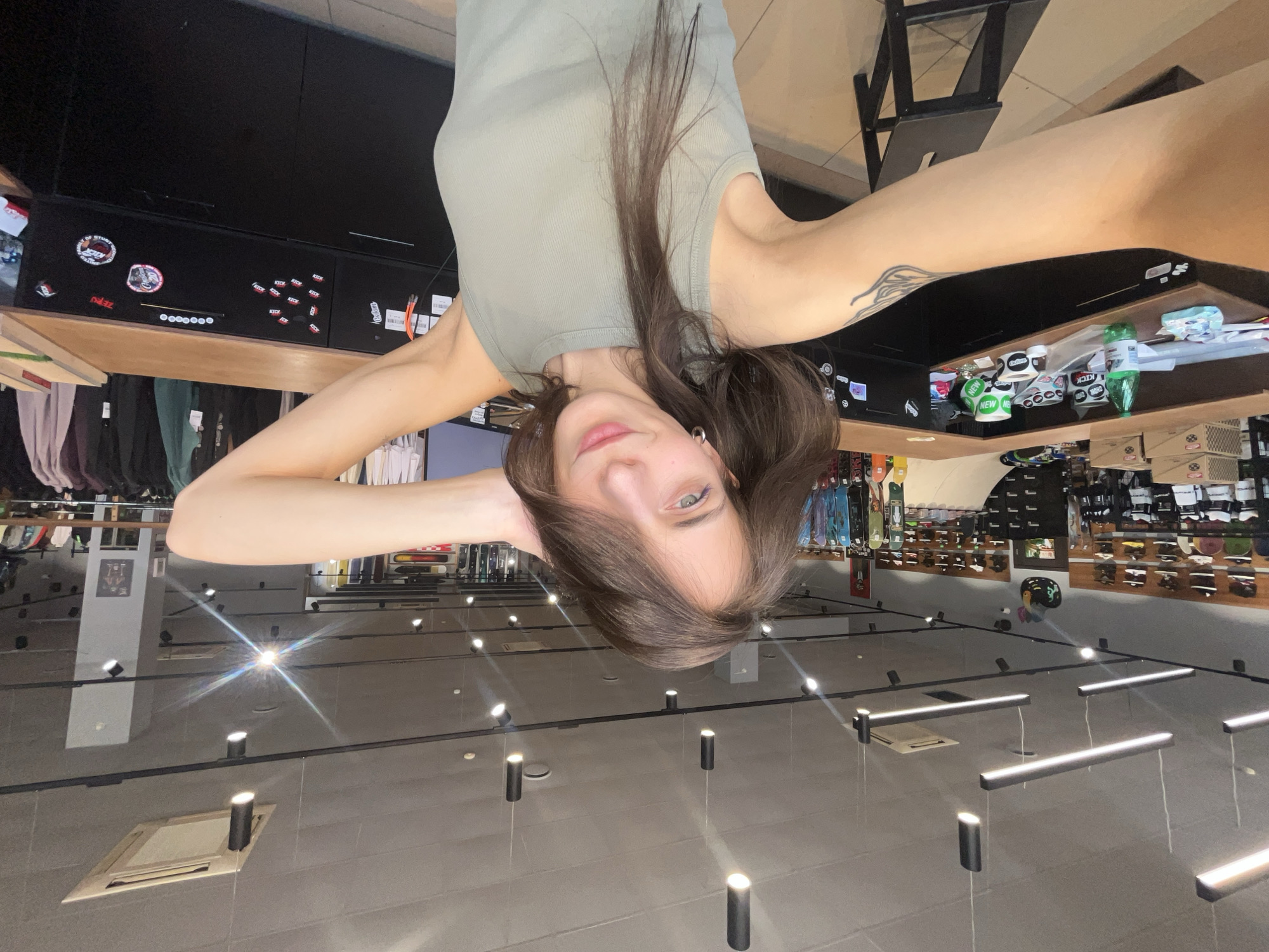 A female is selfieing inside a building. She is wearing a light olive green tank top, and her brown hair is down. A tattoo is on her right arm. There are clothing racks to the left, and shelves with products to the right and behind her. Based on the product branding visible, it is a skateboard shop. The location is in Krasnoyarsk, Russia. Overhead are numerous track lights with rectangular fixtures.