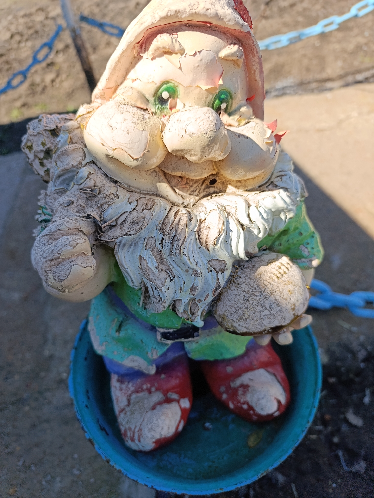 A weathered garden gnome figurine is centered in the image, standing on a blue painted metal object that appears to be a repurposed bucket or similar container. The gnome has a white hat, green eyes, a large nose, and a long white beard. It's wearing a green and purple shirt, and red shoes. The paint on the gnome is peeling and cracked, indicating it has been exposed to the elements for an extended period. The background shows a concrete surface and a blue chain. Based on the request, this scene is likely located in the city of Уральский, Russia.