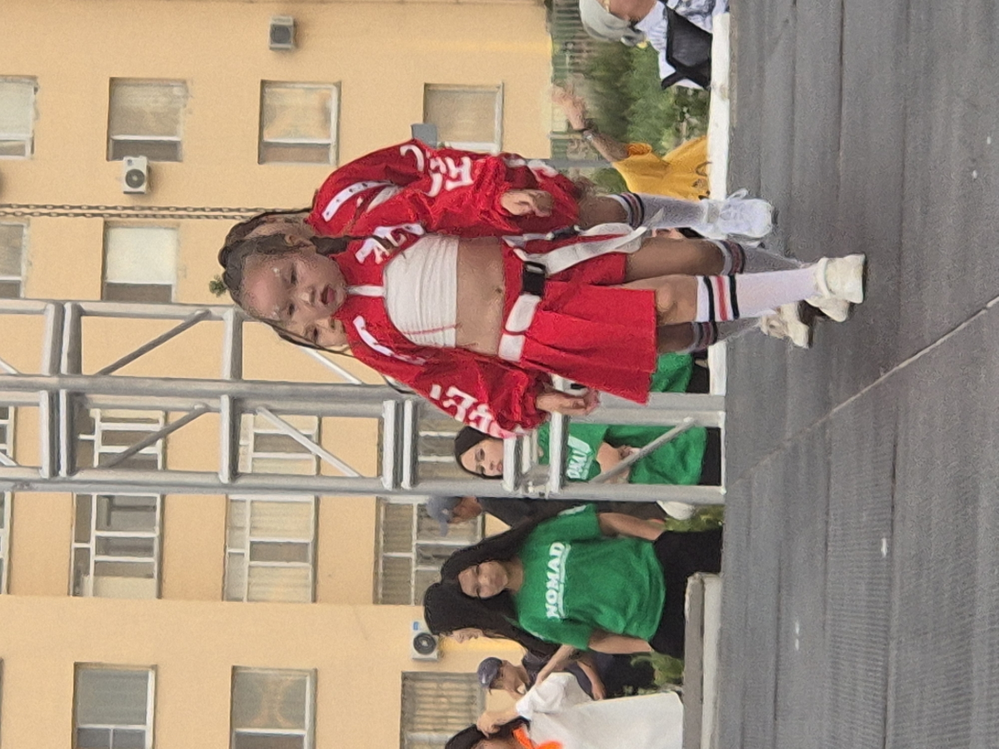 The image captures a group of young people, likely a dance crew, performing outdoors. The central figures are two individuals, one appearing to be a child, dressed in coordinating red outfits with white accents and red skirts. They are wearing white socks with red and black stripes and white sneakers. The child is being held by the other person, who is also dressed similarly. They are on a stage or platform, with a metal scaffolding structure to their left and a grey textured surface beneath their feet, suggesting an outdoor performance area.
In the background, a pale yellow building with multiple windows is visible. The windows have a grid-like structure. There are also glimpses of other individuals in green t-shirts, one of which has the text "NOMAD" printed on it in white. Some people are wearing headscarves. The overall impression is of a lively event, possibly a dance competition or a street performance. The lighting suggests it is daytime, and the weather appears to be mild. The emotions on the faces of the performers are difficult to discern due to the image quality and orientation, but the dynamic poses indicate energetic movement. The presence of the building and the scaffolding suggests an urban or semi-urban setting, consistent with the provided location context of Kokshetau, Kazakhstan.