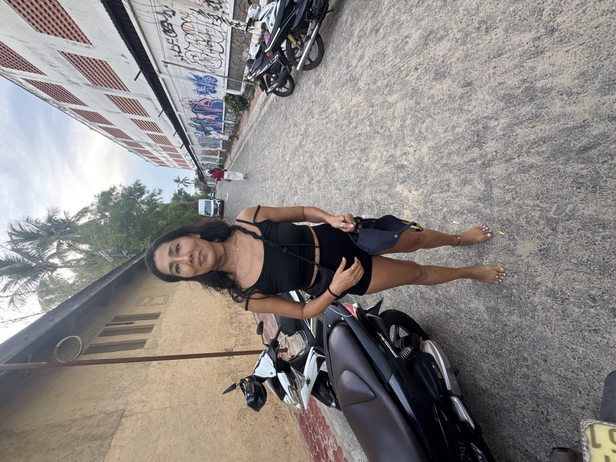 The image shows a woman standing on a street in Mũi Né, Vietnam. She is wearing a black tank top and black shorts. Her toenails are painted white. There are two motorcycles parked behind her. To her left, there is a building with a patterned facade and graffiti on its wall. Further in the background, there are palm trees and a clear sky, suggesting it is daytime. The scene appears to be outdoors on a paved street. The woman is looking towards the camera with a slight smile.