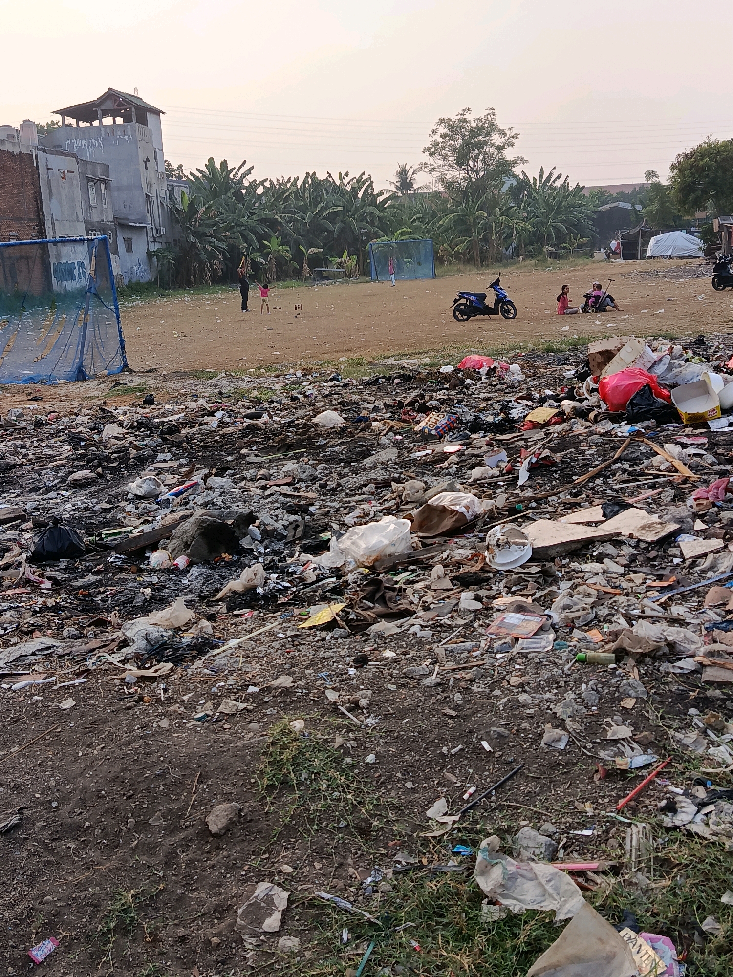The image depicts a vacant lot in a city, likely in Bekasi, Indonesia, laden with debris and refuse. The foreground is dominated by a dense accumulation of garbage, including plastic bags, bottles, cardboard, and what appears to be burnt material, forming a cluttered landscape. In the mid-ground, a sandy or dirt field stretches out, with some sparse vegetation and scattered pieces of trash. Further back, several people, including children, are visible in the field. Two adults and two children appear to be playing or engaged in an activity near some wooden posts. Several motorcycles are parked in this area. In the background, there are buildings with a distinctive architectural style, including a multi-story structure with an elevated section. A cluster of banana trees and other foliage separates the field from more distant structures, including a tent-like shelter and what appear to be informal settlements. The sky is overcast with a pale hue, suggesting it may be early morning or late afternoon.