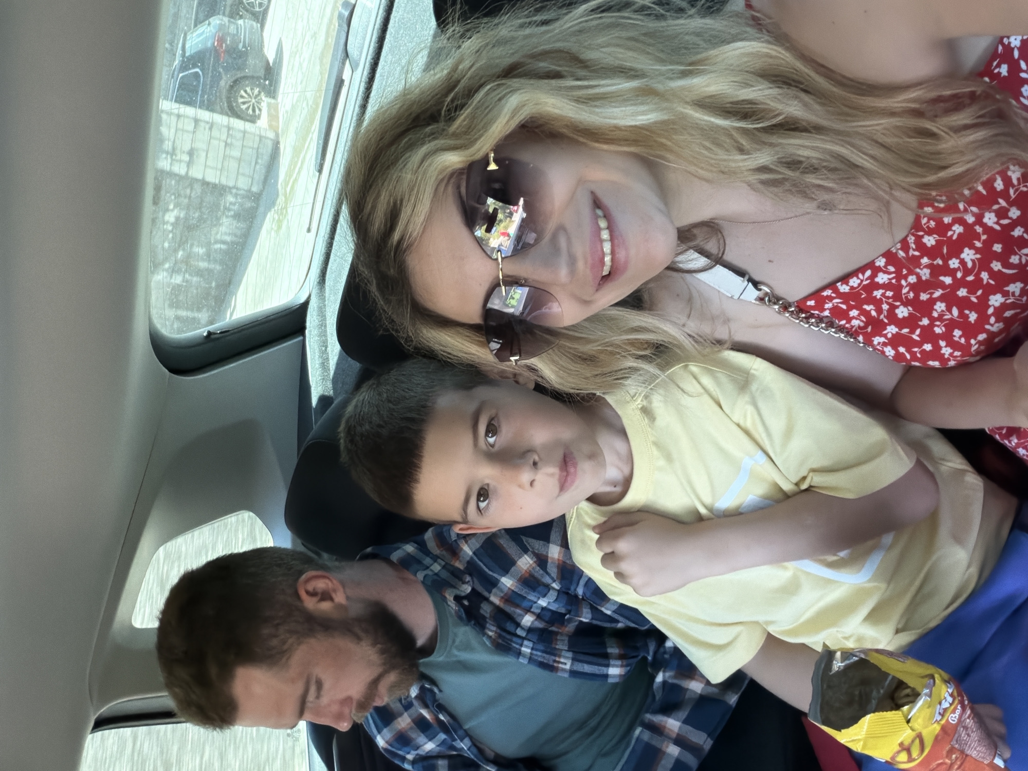 The image depicts three individuals in the back of a vehicle. A woman with long blonde hair and sunglasses is in the upper right, smiling. A young boy wearing a yellow shirt is beside her, holding a bag of pretzels. A man with a beard is in the lower left, looking down. Outside the vehicle, the reflection in the car window shows a cityscape and another car. Given the request for location specifics, the city of Dobrota, Montenegro is mentioned, but cannot be verified from this image alone.